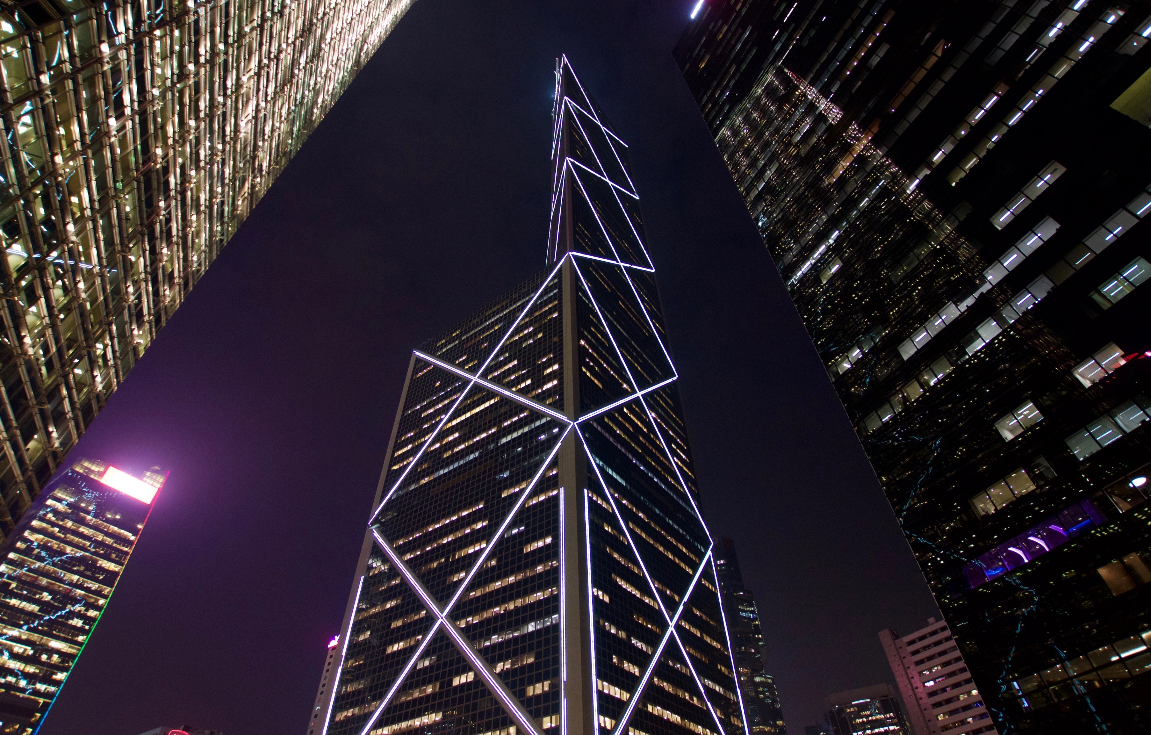 A brightly illuminated angular skyscraper rises above surrounding buildings at night.
