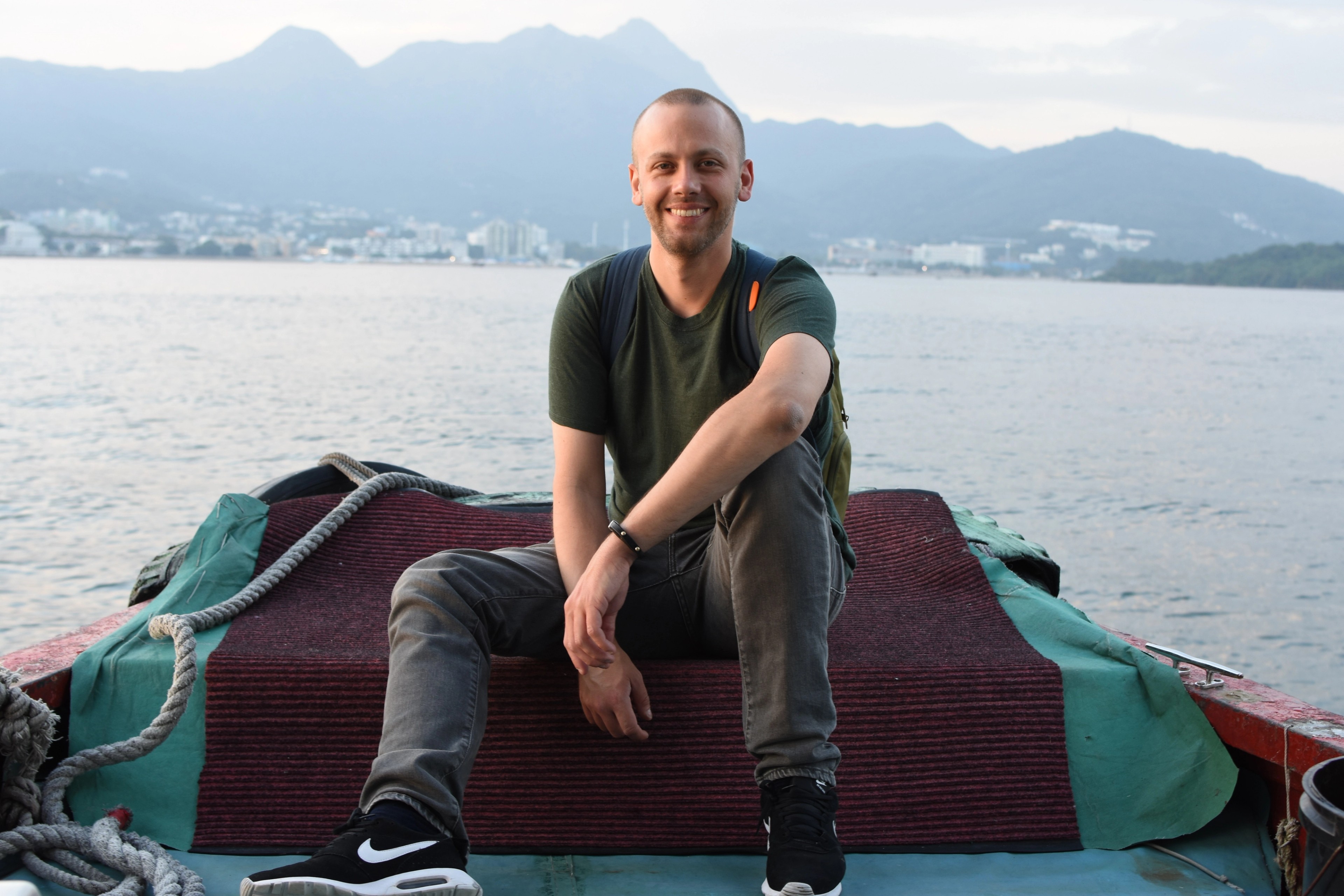 A smiling man sits on the bow of a small boat with mountains behind him.