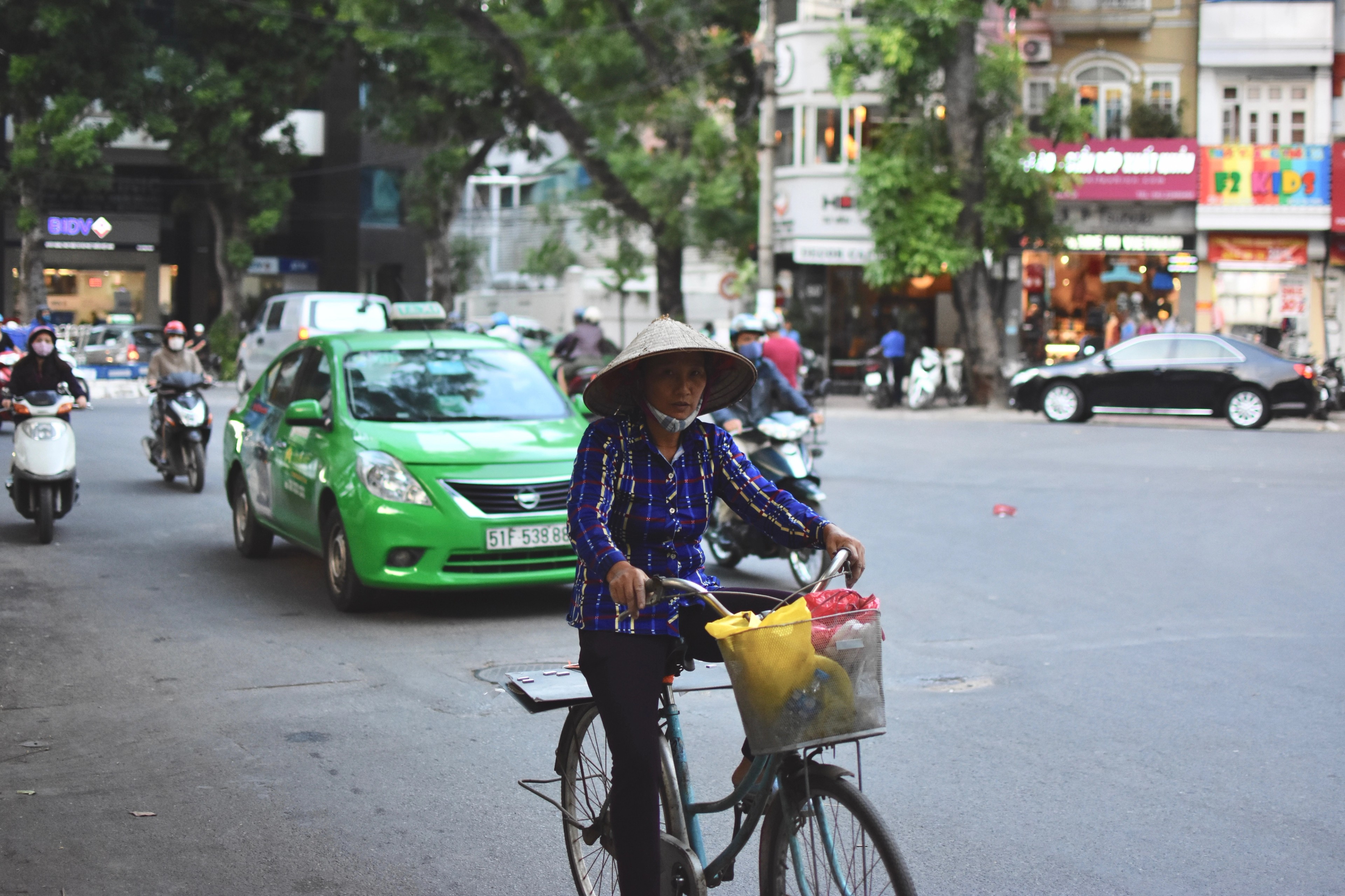 A flower vendor on a bicycle passes a green taxi at an intersection.