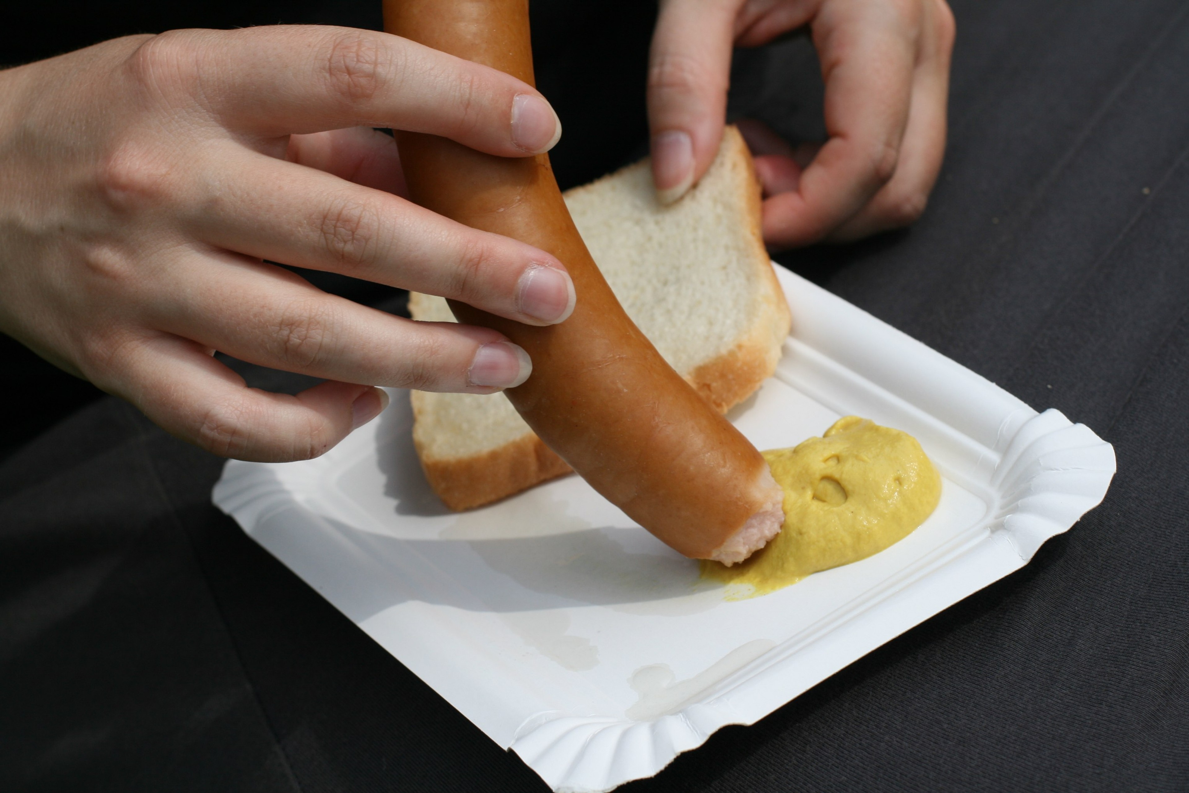 Hands hold a bread roll above a paper tray with mustard.
