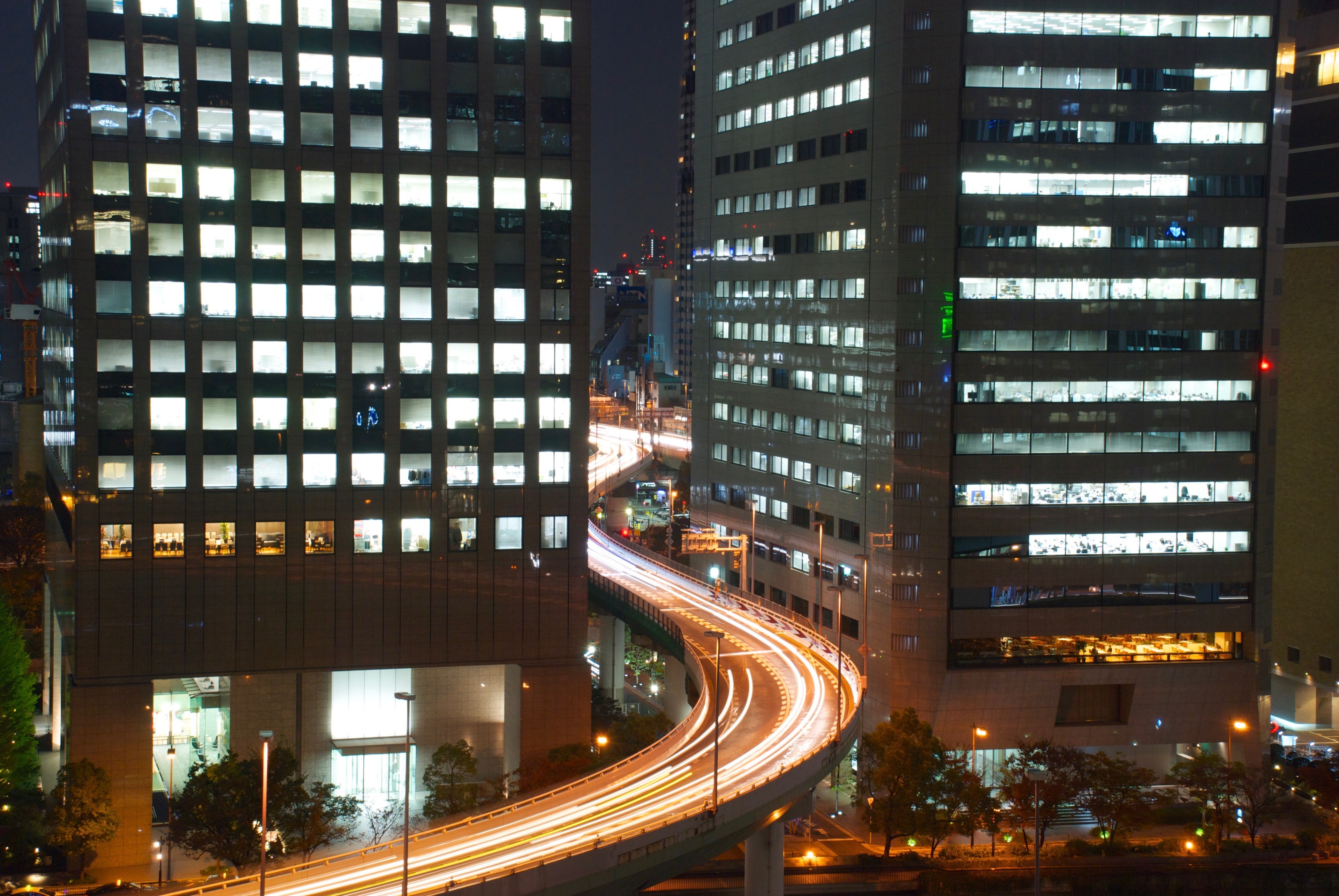 Curving vehicle lights pass between illuminated office buildings at night.
