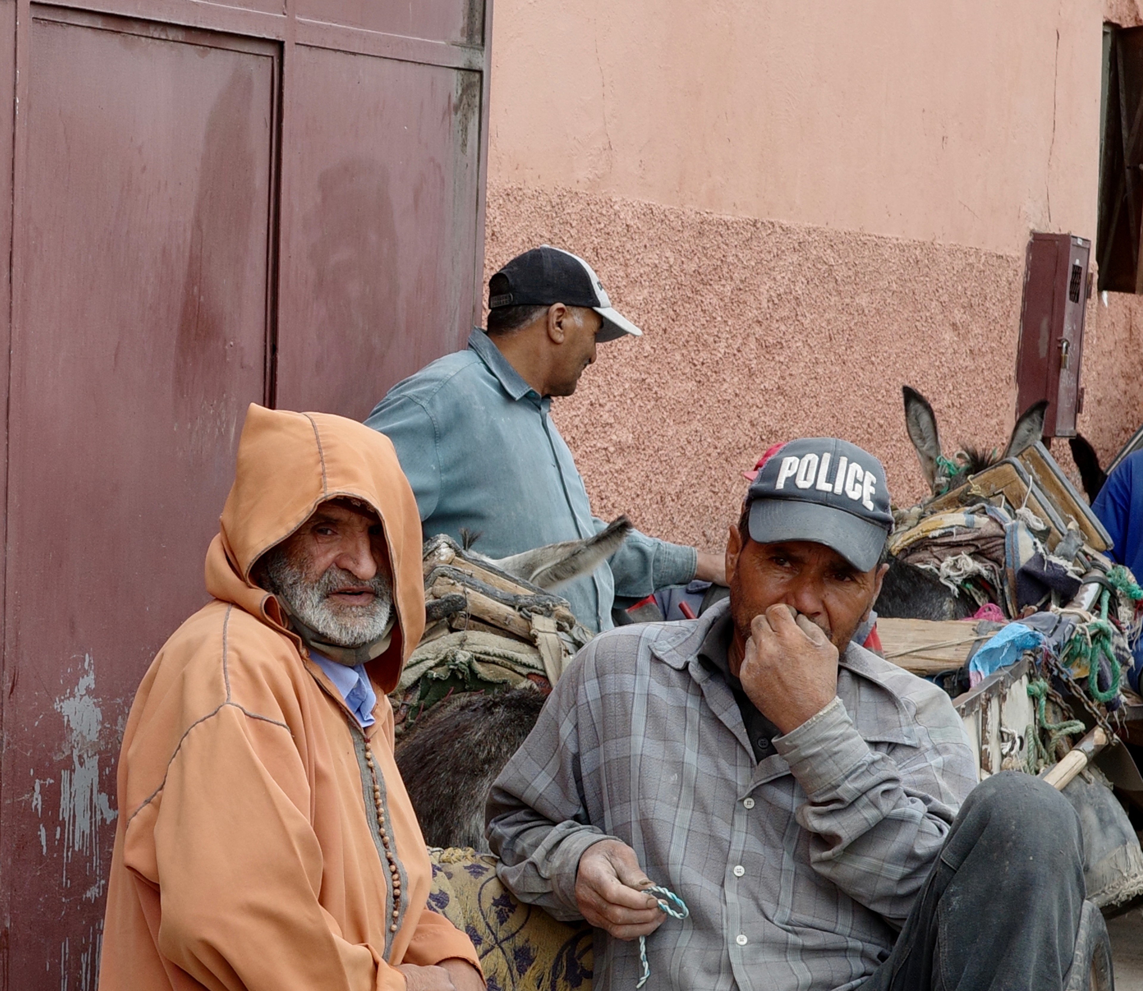 Two men in hats sit beside piles of materials against a pink wall.