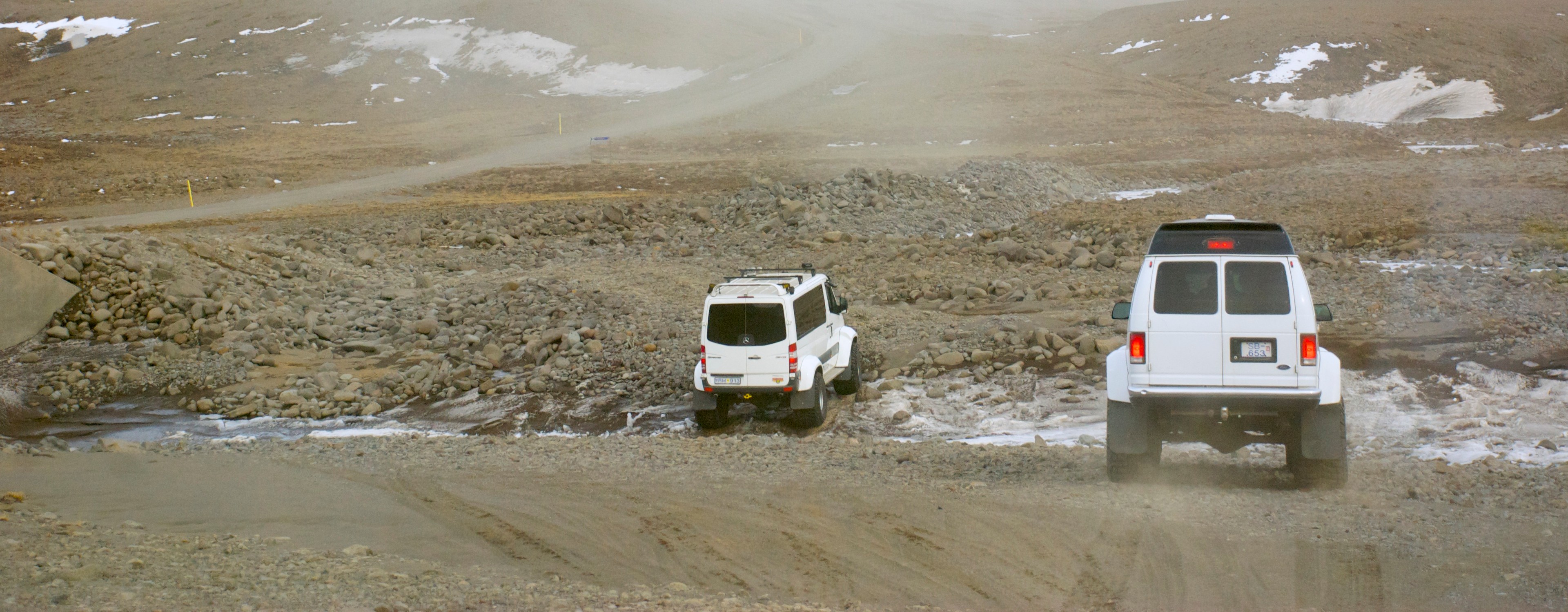 Two white vehicles drive across a flat, stony landscape threaded with water.