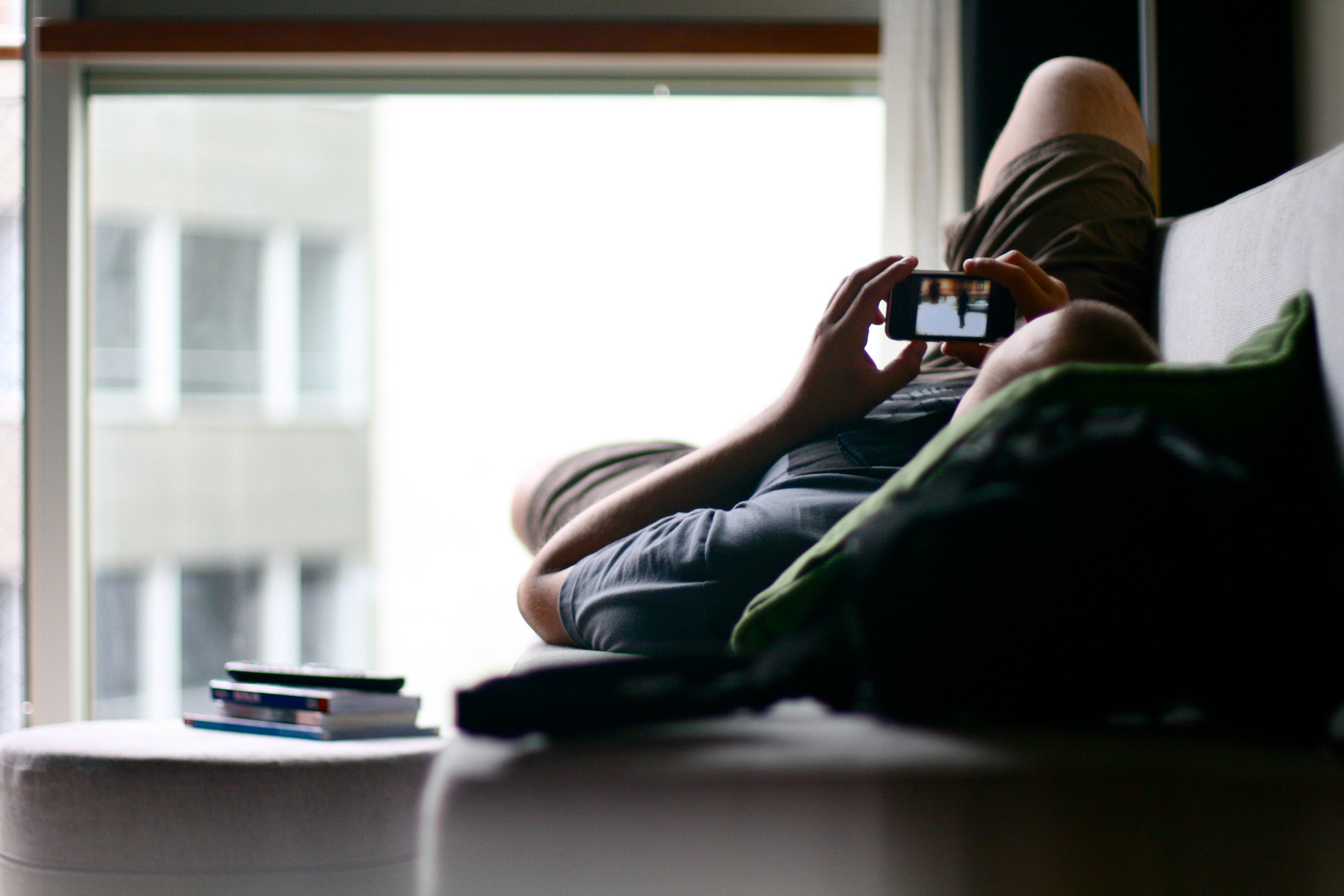 A person sits on a window ledge using a phone, seen in silhouette.