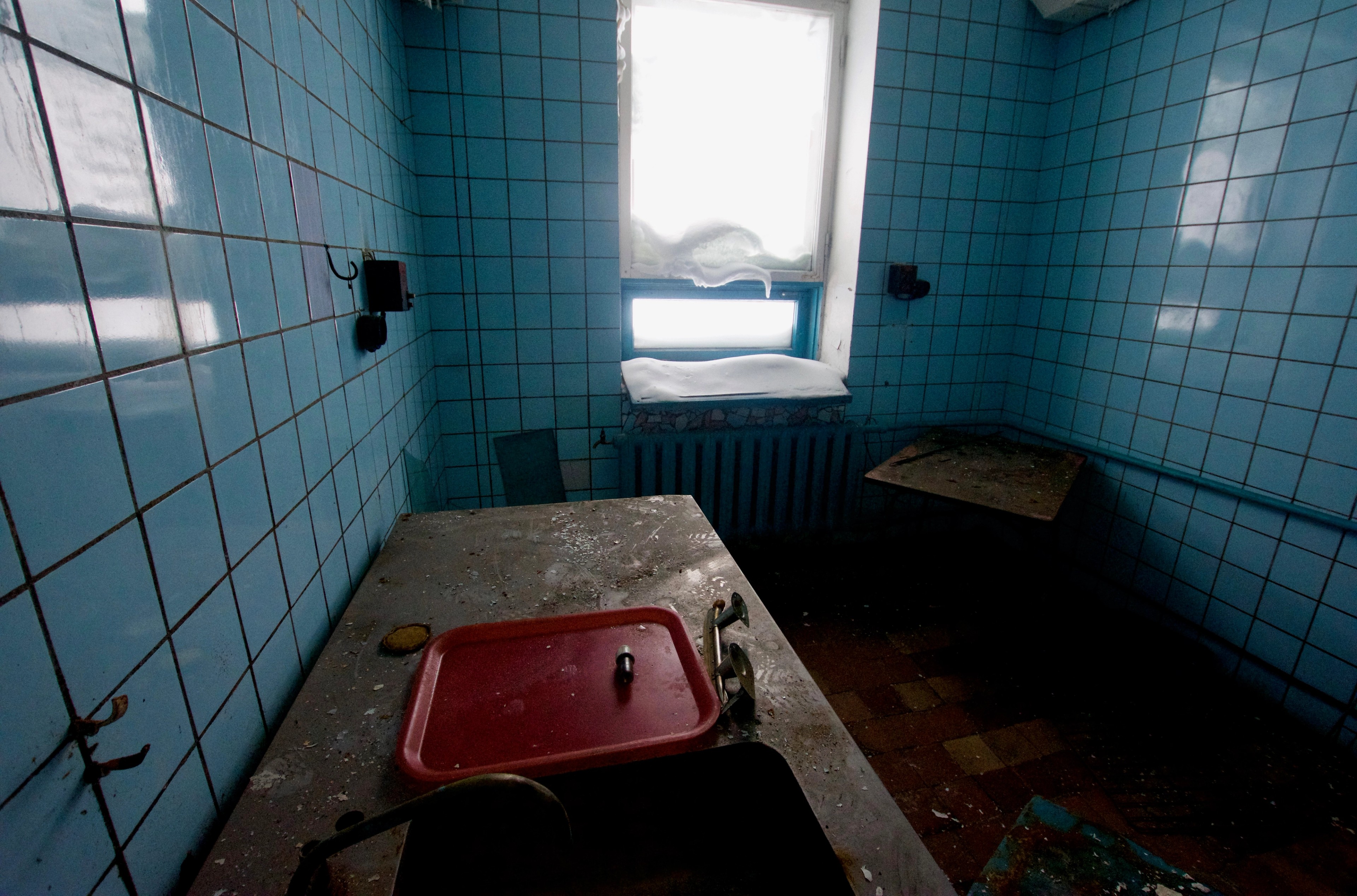 A worn washbasin sits below a window in a tiled room.