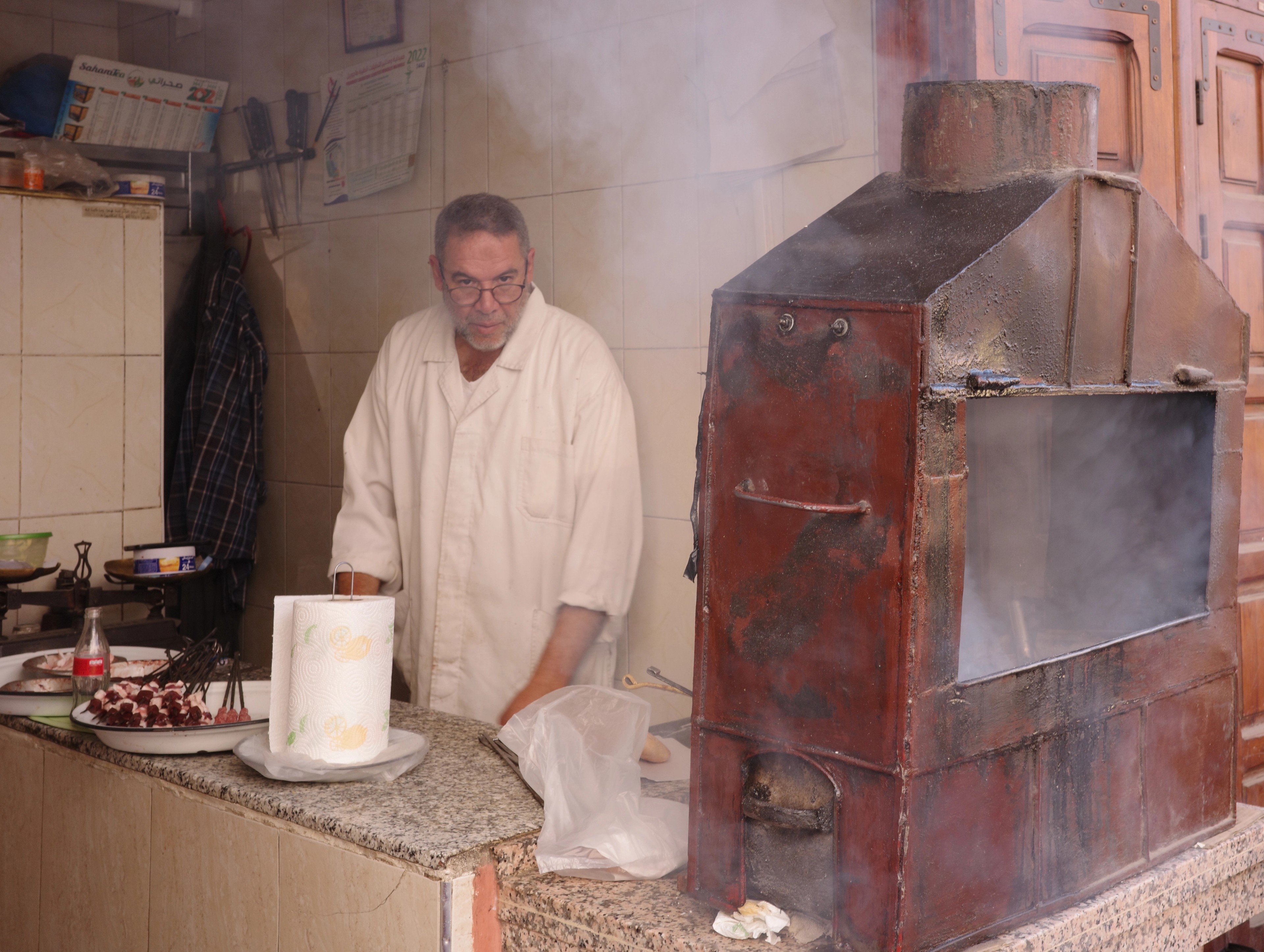 An older man in a white robe works at a counter in a tiled kitchen.