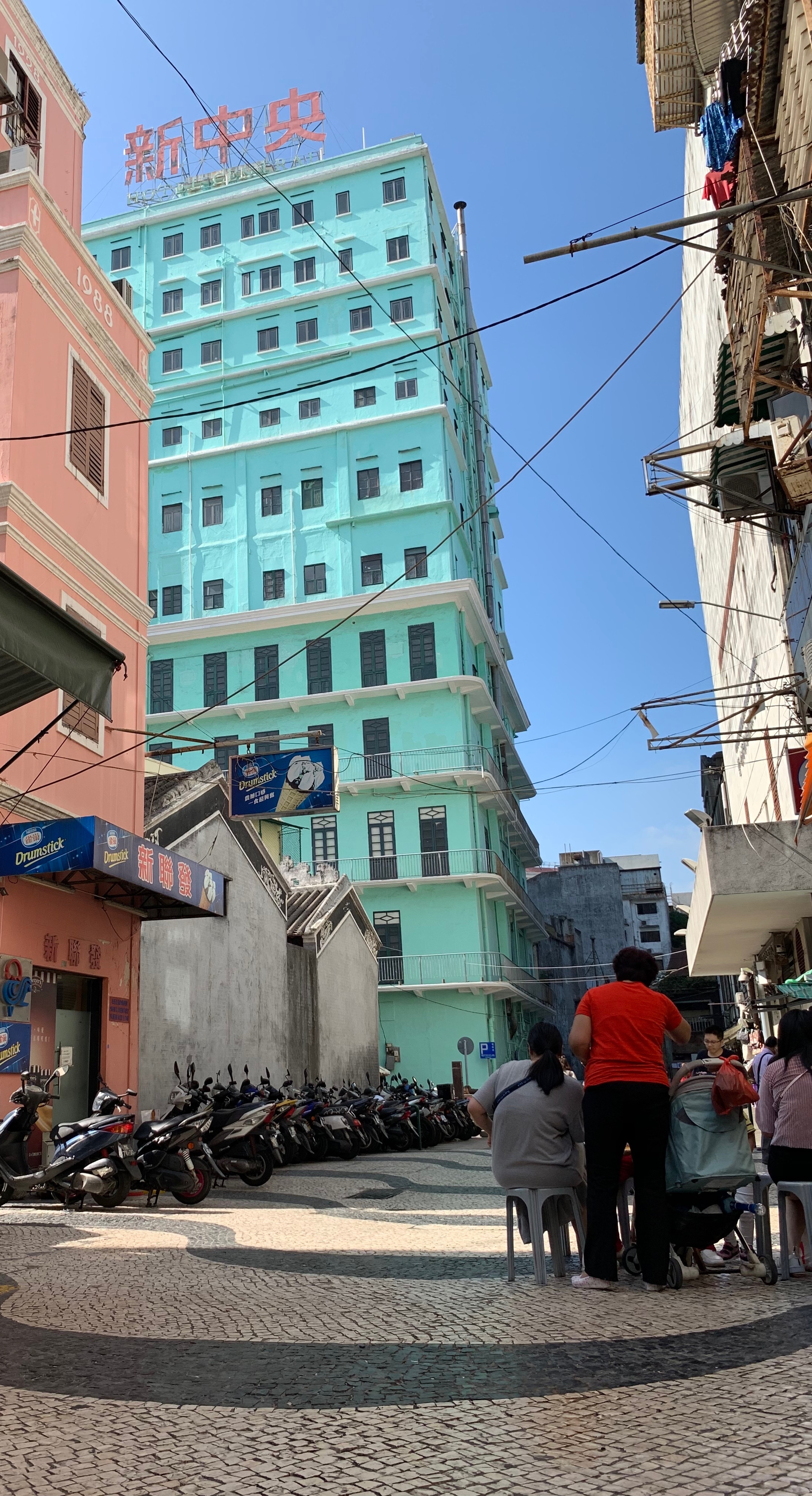 People walking beside a turquoise building on a narrow Macau street.