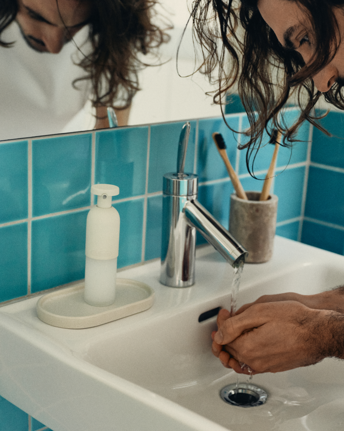 A person washes their hands at a bathroom sink, using a stream of water from a silver faucet. A soap dispenser, toothbrush holder, and soap dish are also present on the sink.