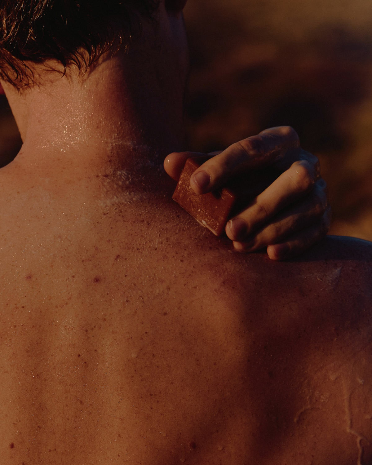 A person applies a reddish-brown bar of soap to their back, visible on their skin.