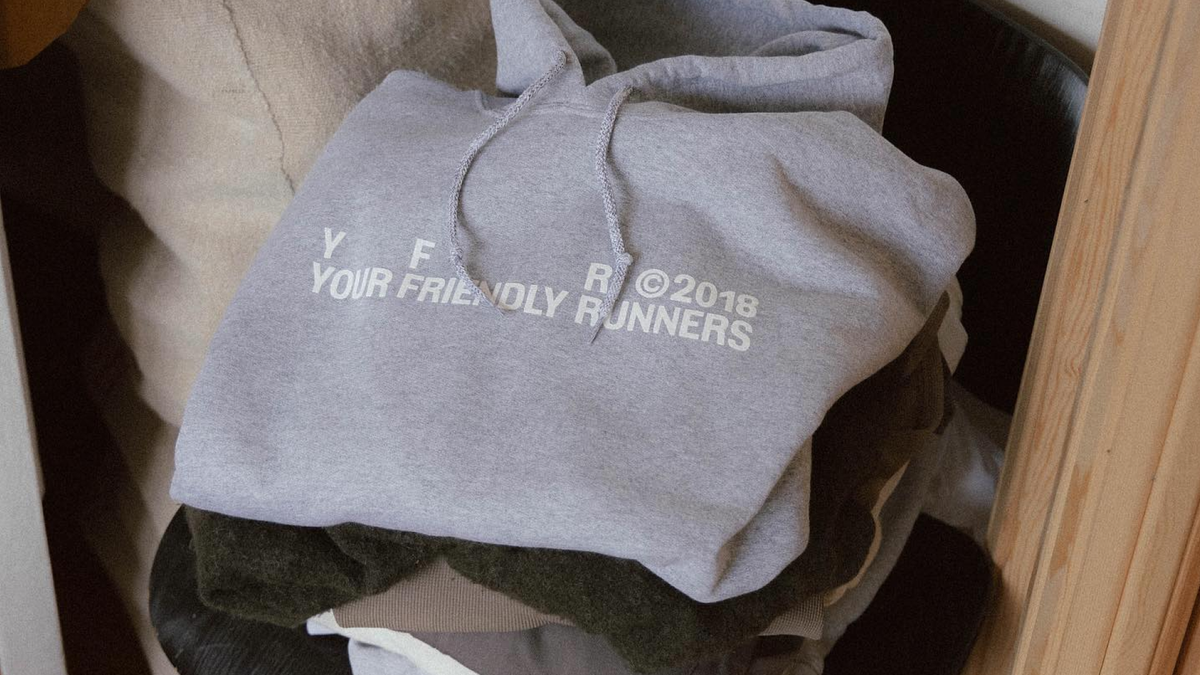 A gray hoodie with "YF FRIENDLY RUNNERS" and "R ©2018" printed in white lies stacked on a pile of folded clothing.
