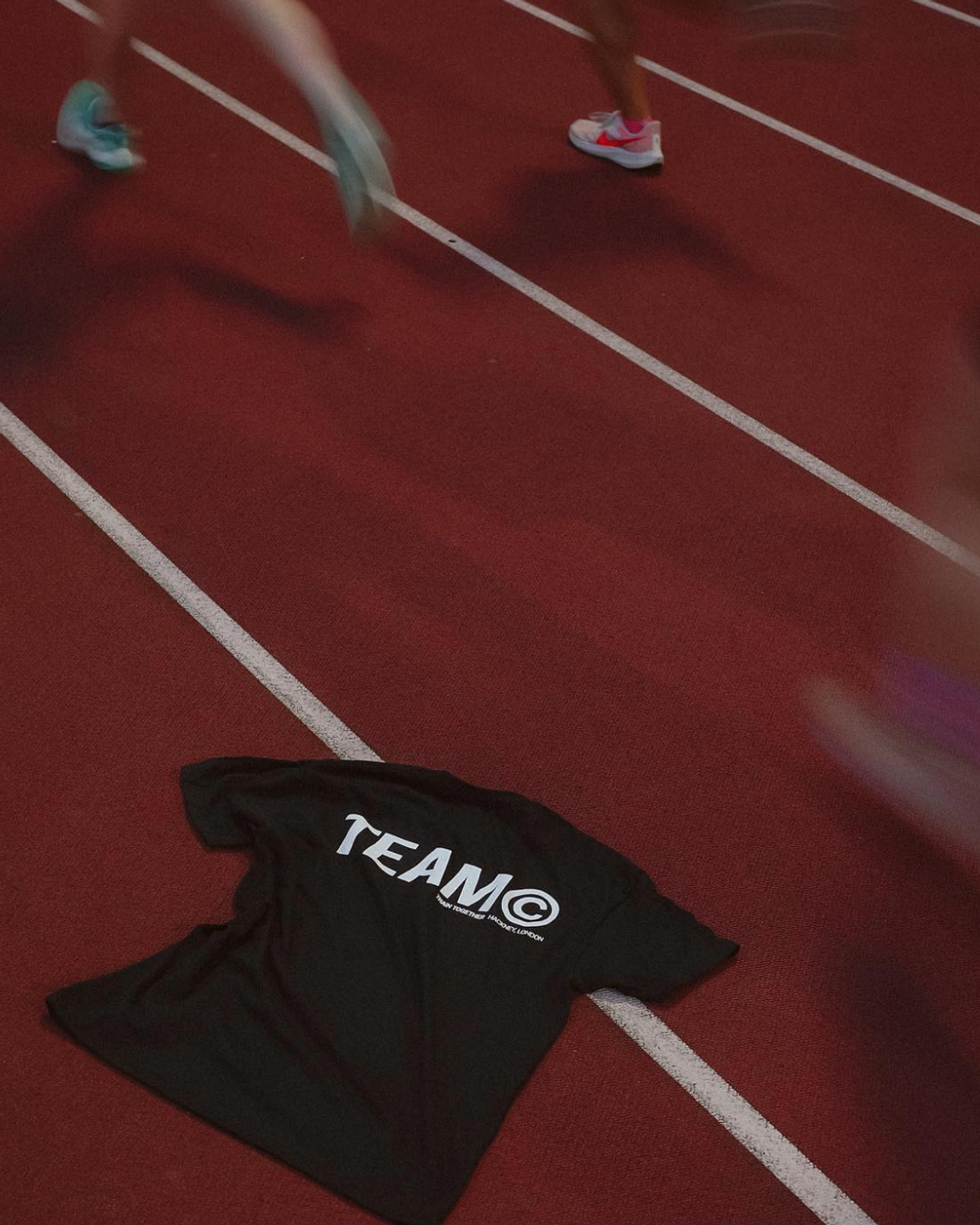A black t-shirt with "TEAM" and a registered trademark symbol lies on a running track. Blurred figures in athletic shoes are visible in the background.