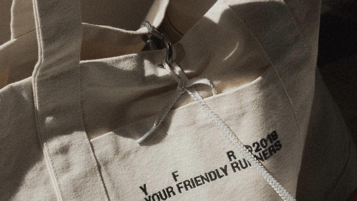 A beige canvas tote bag with black embroidered text reads "YOUR FRIENDLY RUNERS 2018". A white drawstring is tied through the top opening.
