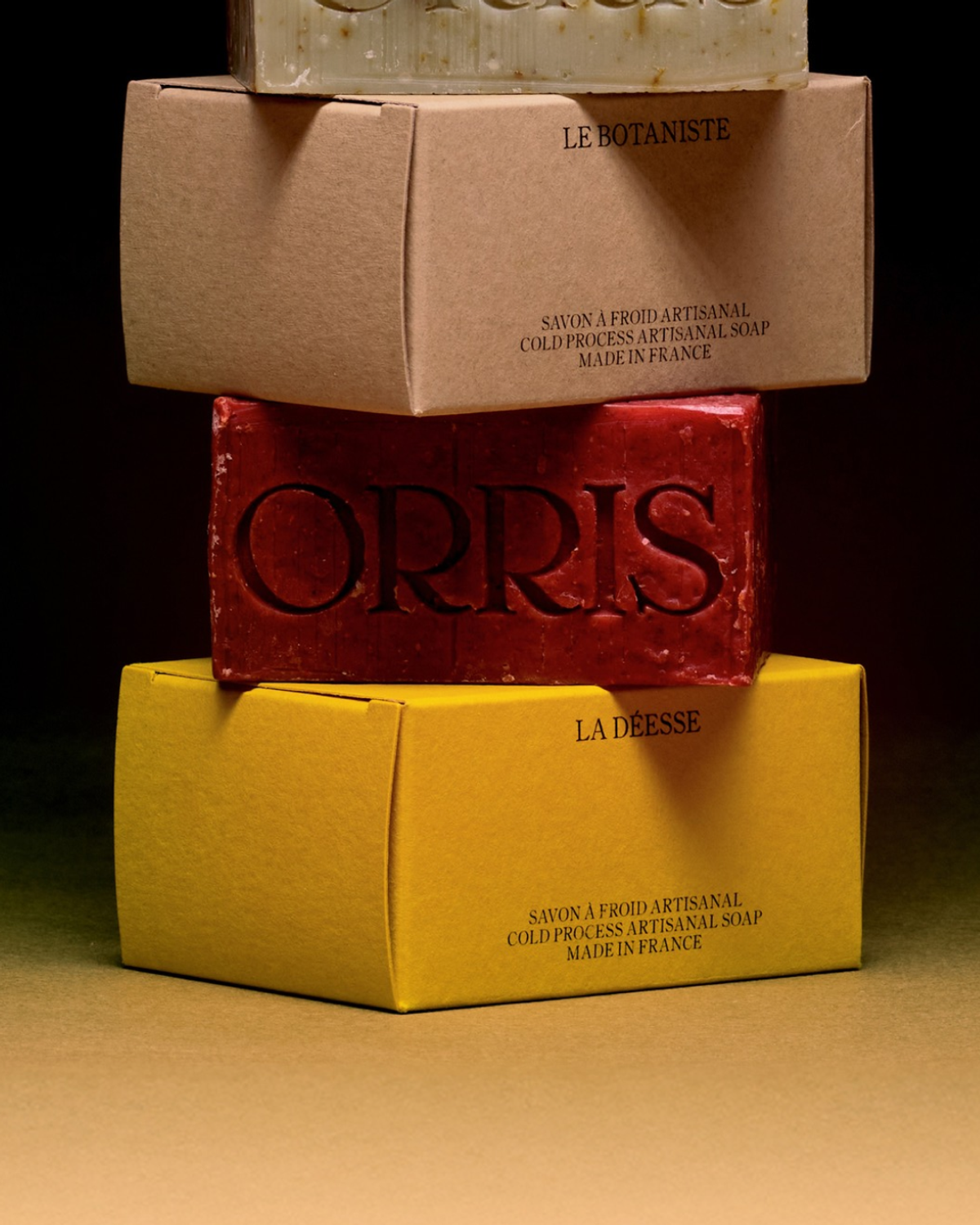 Three boxes of artisanal soap are stacked. The top box is light beige, the middle is dark red with "ORRIS," and the bottom is yellow with "LA DEESSE." Each box indicates "SAVON A FROID ARTISANAL" a...