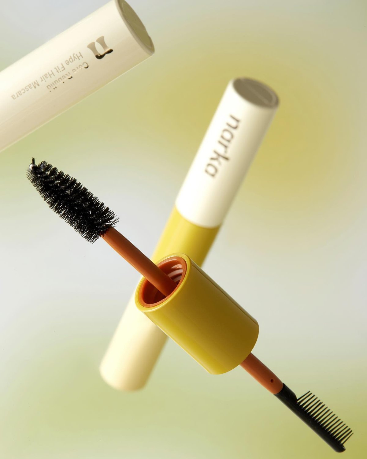 Two Marka mascara tubes, one open with a brush and wand, are shown against a light green background. The tubes are off-white with yellow accents and feature the brand name.