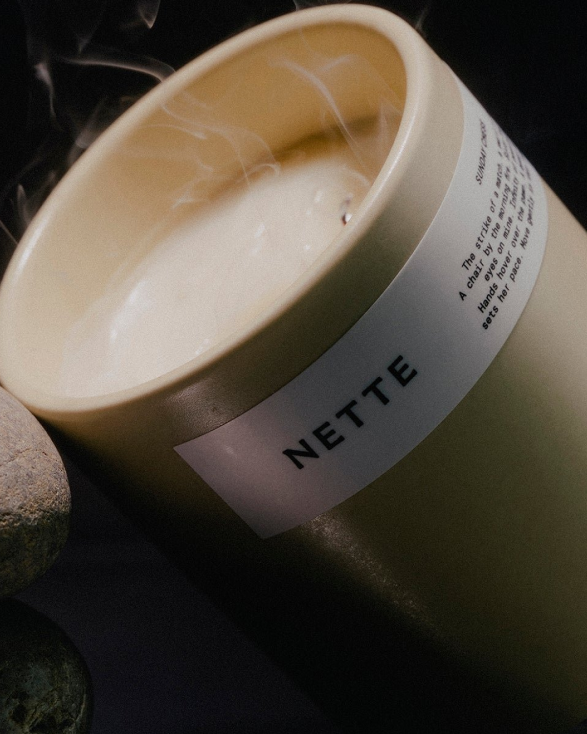 Beige NETTE candle with smoke rising from wick on dark surface