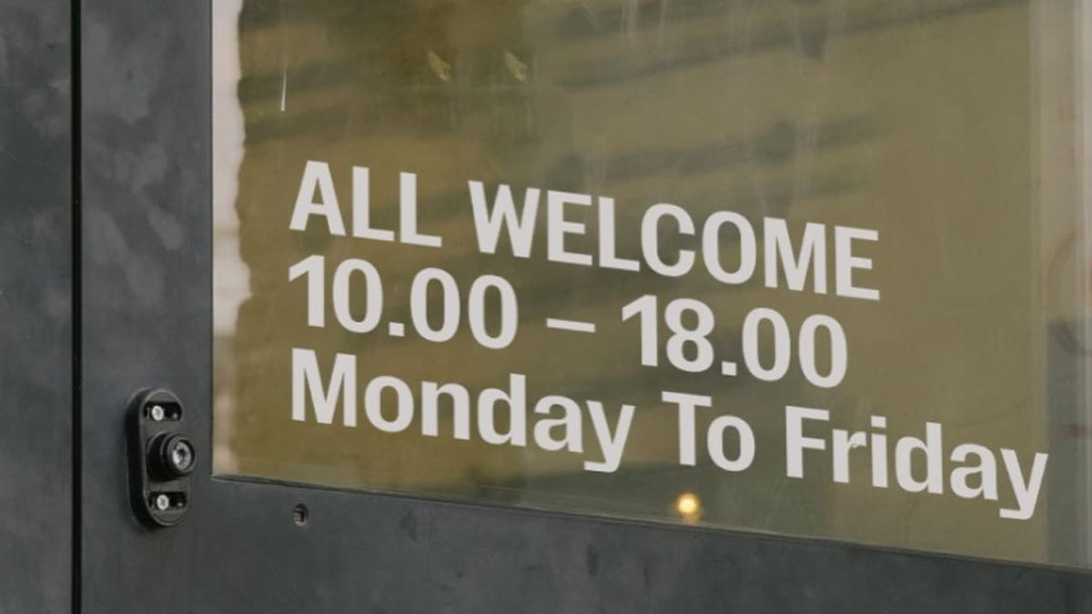 A glass door with a "ALL WELCOME 10:00 –18:00 Monday To Friday" sign. The door is dark gray or black and has a keyhole.