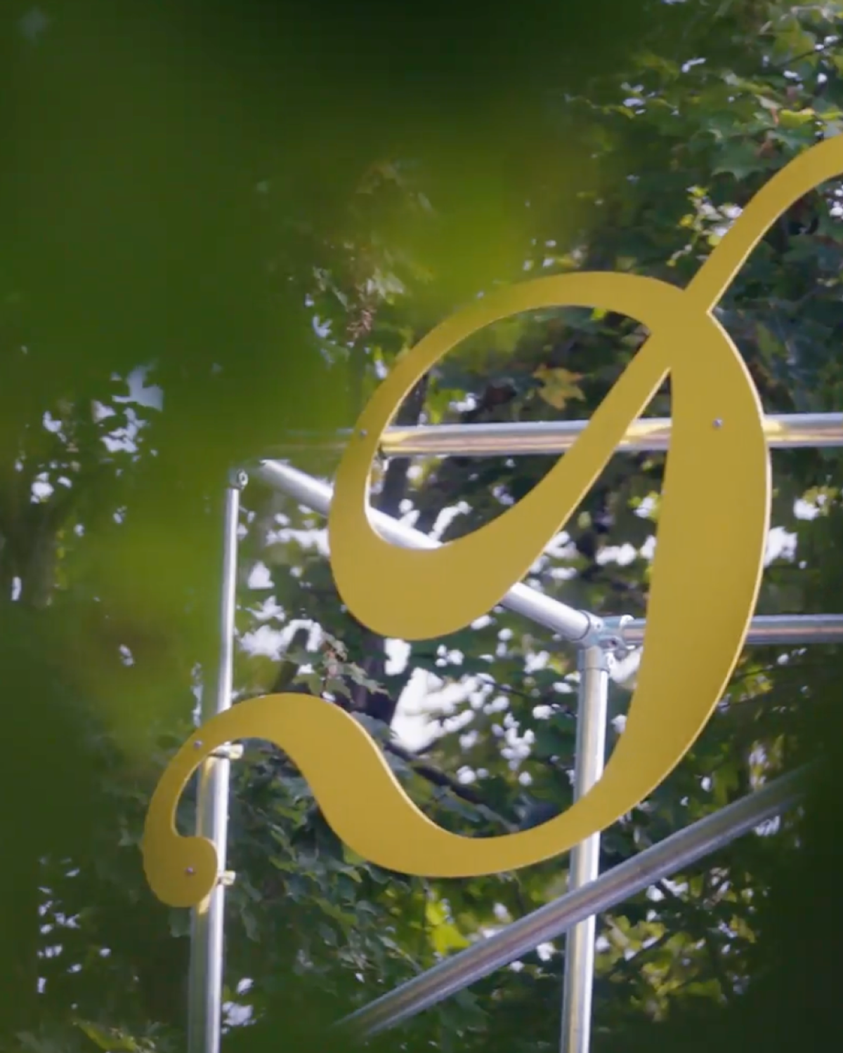A large, stylized yellow letter "P" on a metal framework is partially obscured by green foliage.