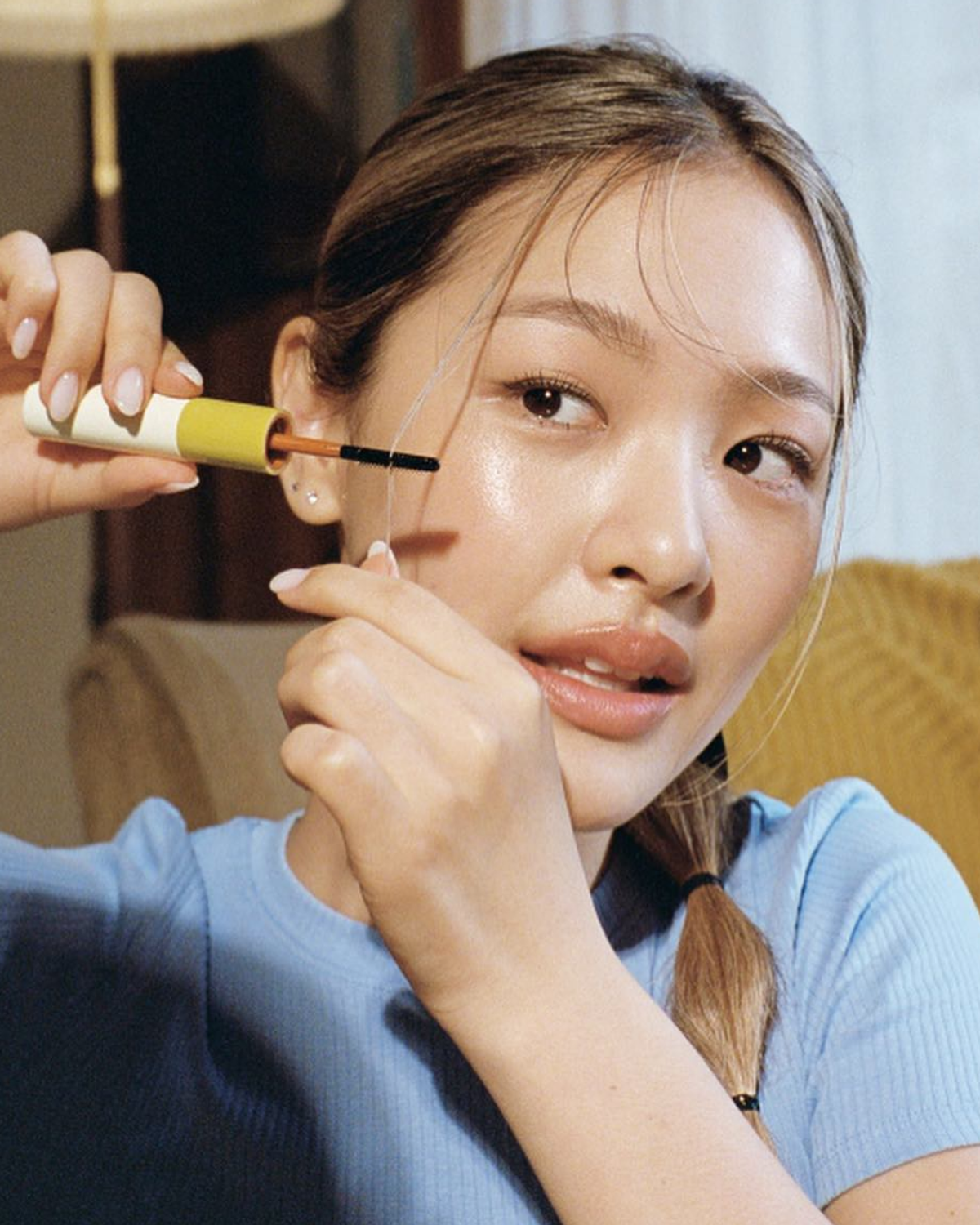 A young woman applies mascara with a yellow tube to a thin black line on her eyebrow. She wears a light blue top and has her hair tied back in a braid. A lamp and patterned fabric are visible in th...
