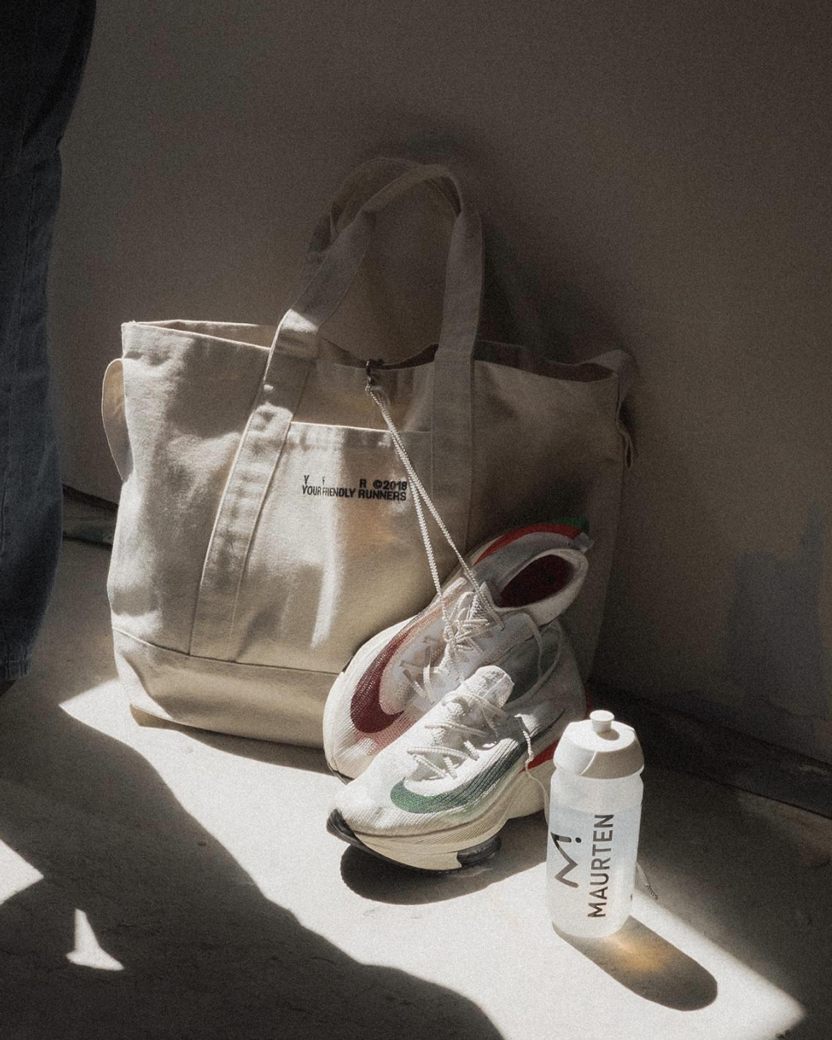 A beige tote bag labeled "YOUR FRIENDLY RUNERS," a pair of white running shoes, and a white water bottle labeled "MAURTEN" are arranged on a light-colored floor.