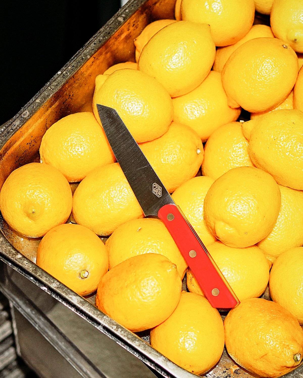 A metal tray holds a collection of whole lemons. A serrated knife with a red handle rests on top of the lemons.