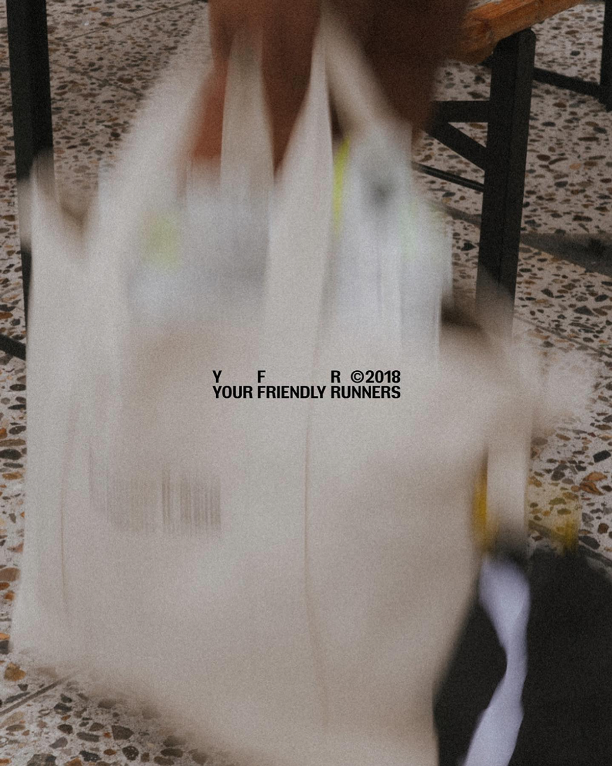 A person holds a white tote bag with the text "Y F F 2018" and "YOUR FRIENDLY RUNNERS" superimposed. The background is blurred, showing a tiled floor and dark objects.