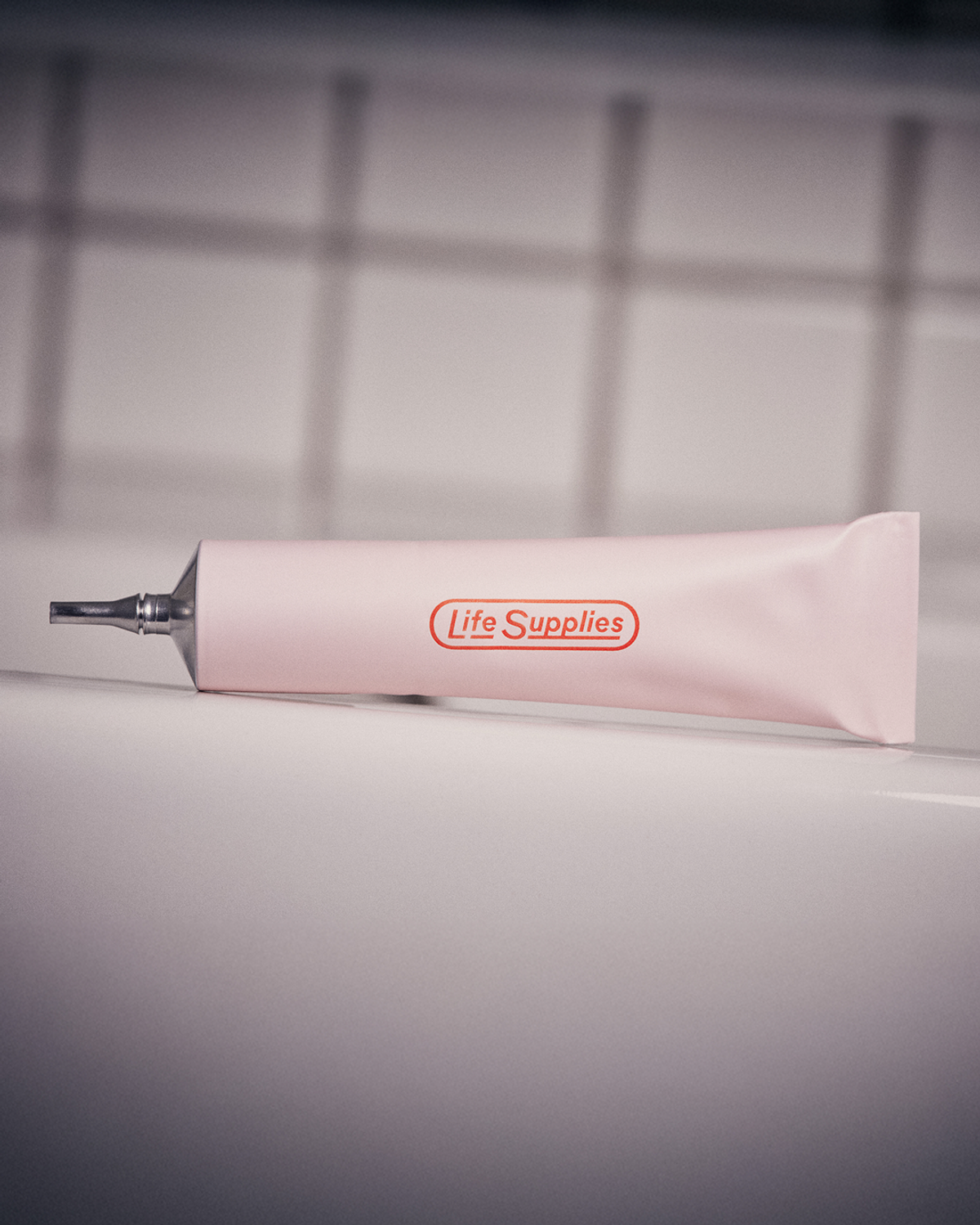 A pale pink tube of Life Supplies product rests on a white tiled surface. The tube has a silver metal tip and features the brand name in red.