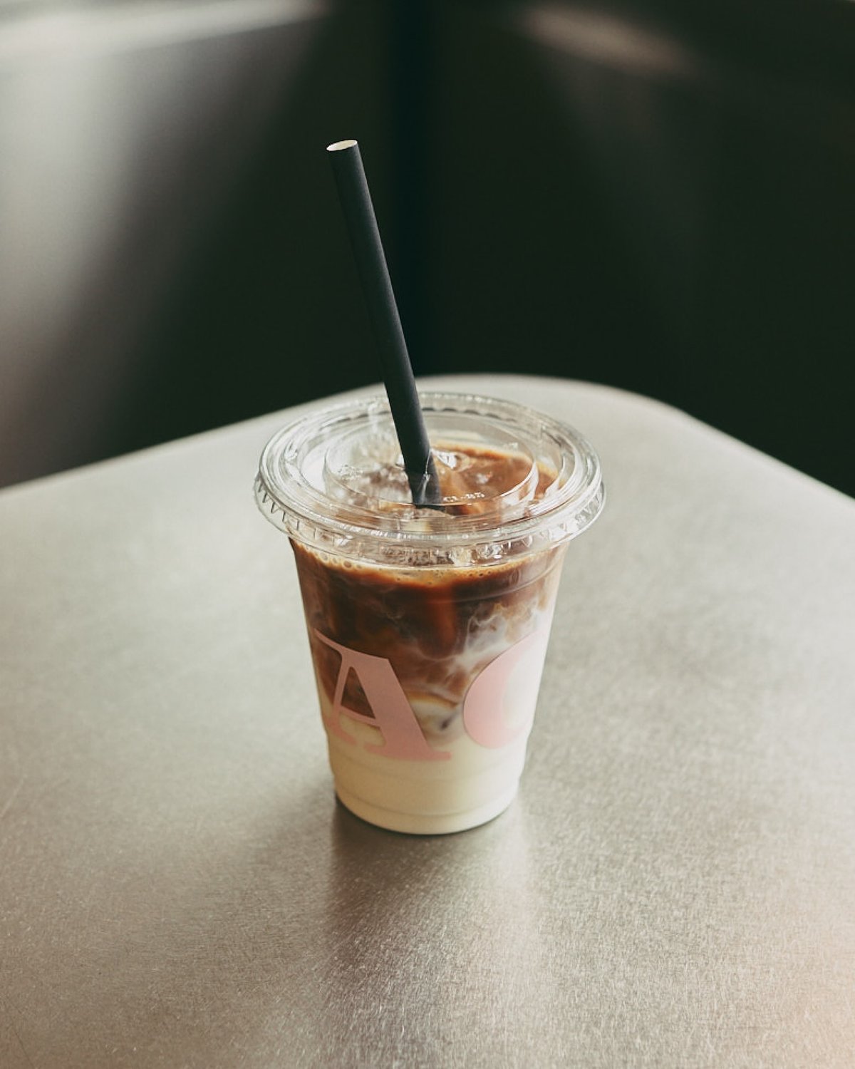 A clear plastic cup containing iced coffee with milk and a black straw sits on a metallic surface. The cup features the letters "AC" in pink.