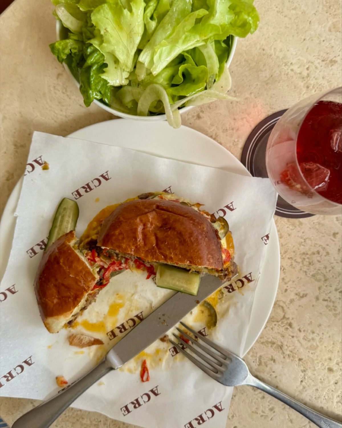 A partially eaten burger with lettuce, pickles, and sauce sits on a white plate, accompanied by a fork and knife. A bowl of salad and a glass of red liquid are also present.