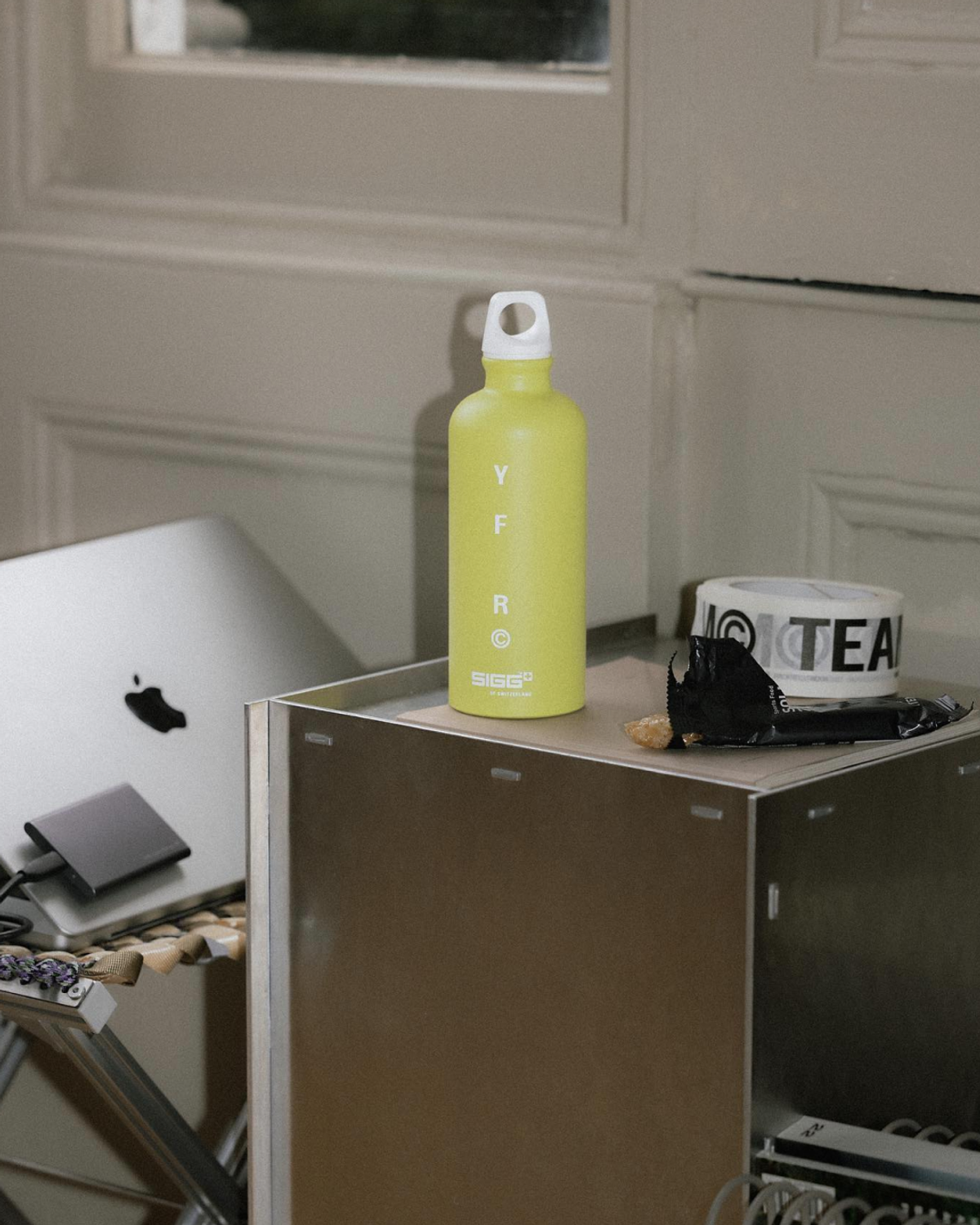 A light green water bottle labeled "YF R" sits on a metallic surface next to a closed laptop and a roll of tape labeled "TEA."