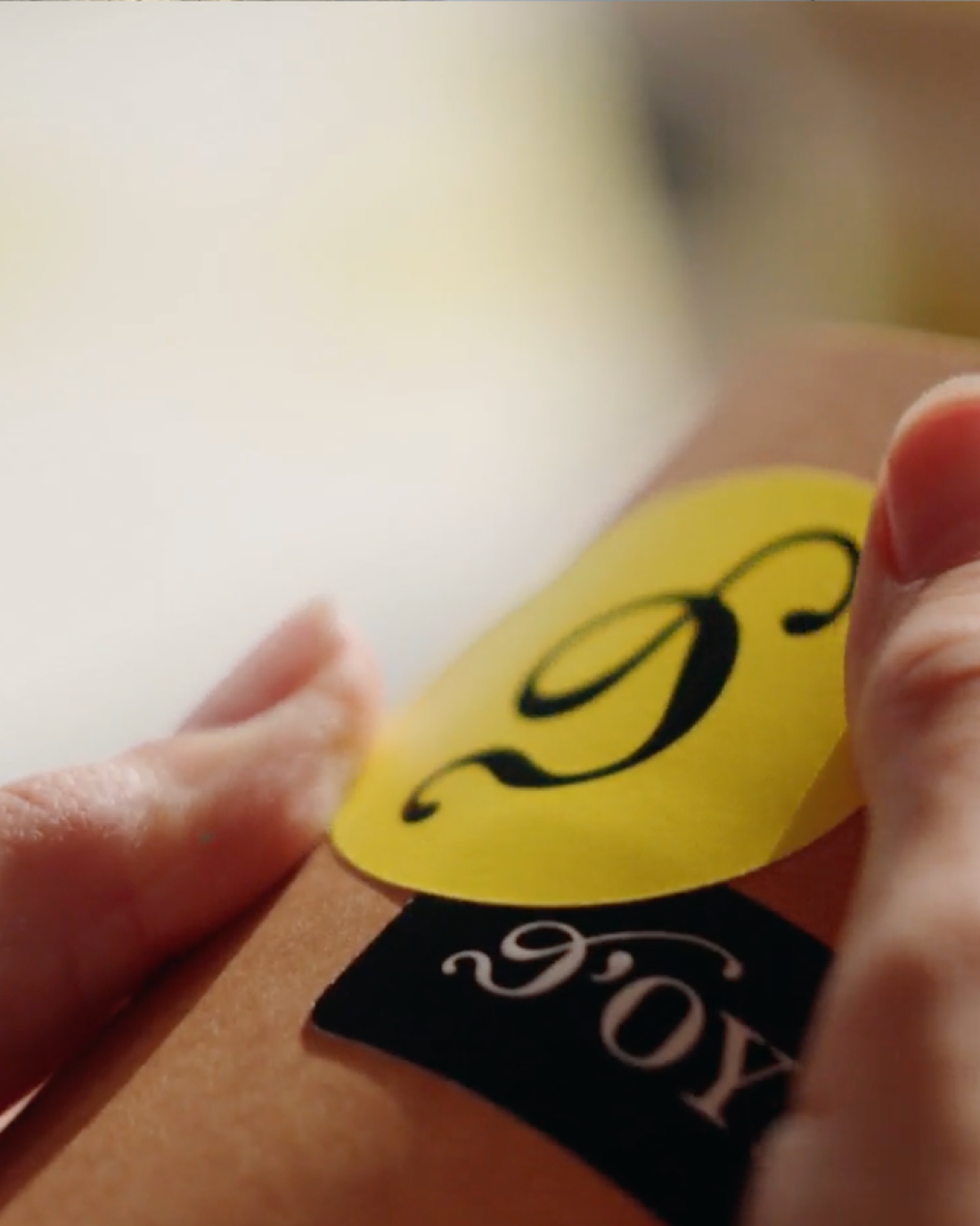 A hand holds a brown surface with two stickers. One sticker is yellow with a black stylized "D," and the other is black with white cursive lettering partially visible.