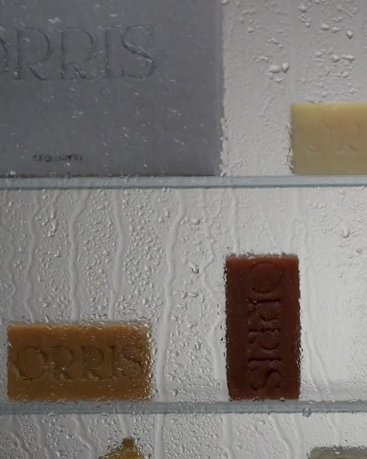 Three rectangular bars of soap, labeled "ORRIS," are displayed on a glass shelf. Water droplets are visible on the shelf's surface.