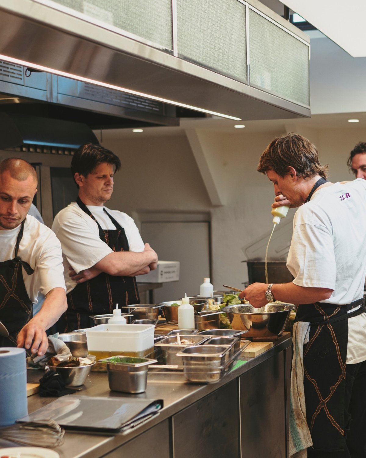 Chefs work in a commercial kitchen, preparing food at a stainless steel counter. Various ingredients, containers, and utensils are visible.