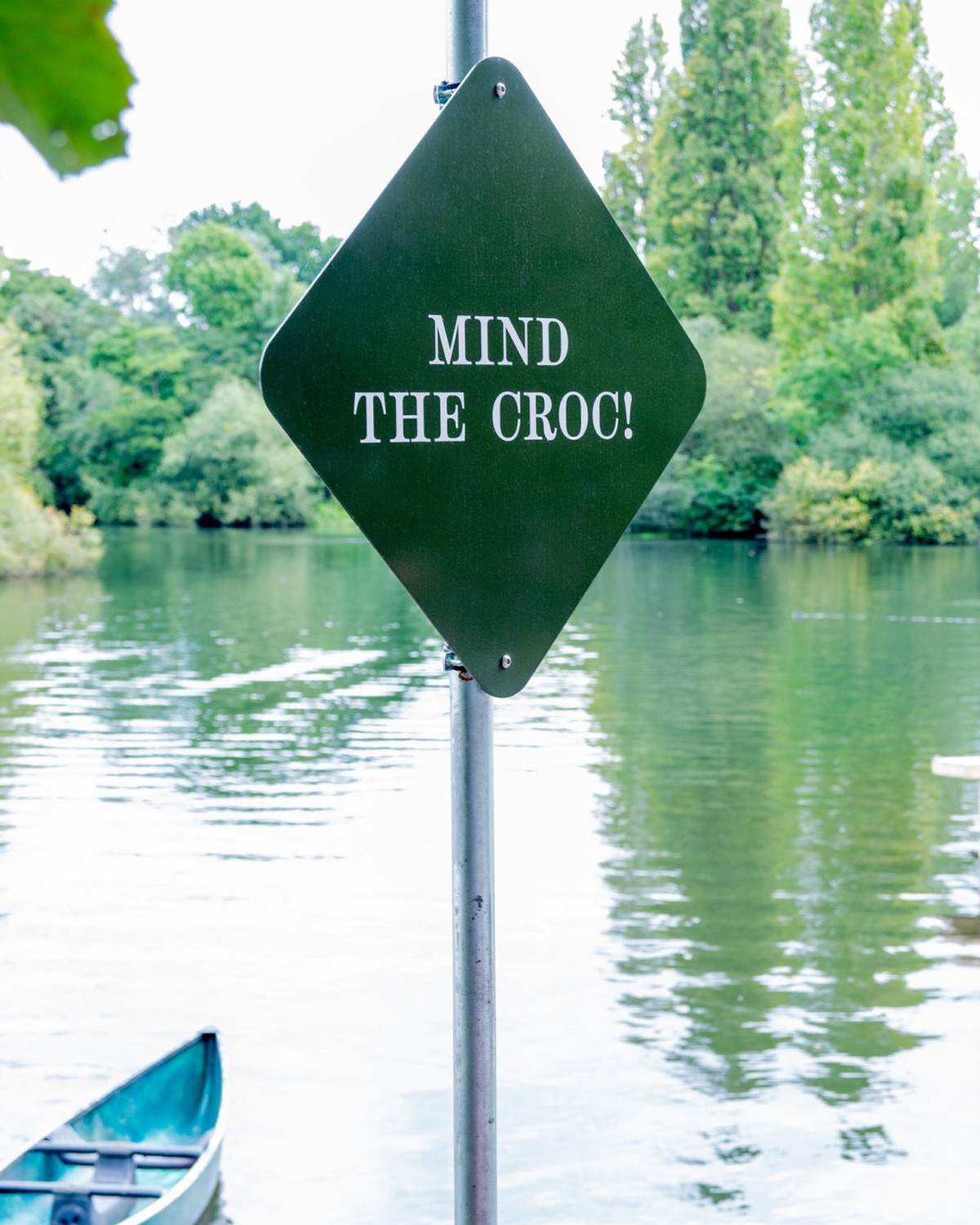 A green diamond-shaped sign reading "MIND THE CROC!" stands on a metal pole beside a calm body of water. A portion of a green canoe is visible in the lower left corner.