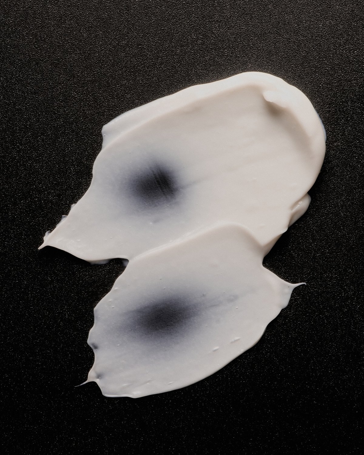 Two swatches of cream or lotion are shown against a black background. The swatches exhibit a creamy texture with darker circular patches.