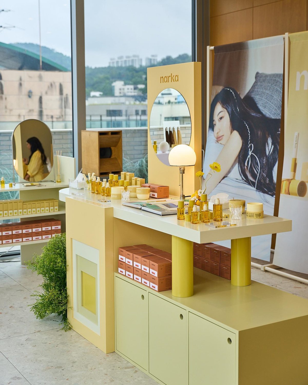 A Narka display featuring a pale yellow countertop with perfume bottles, jars, and a lamp. A large poster of a woman is positioned behind the counter. Boxes labeled "Narka" are stacked on the count...