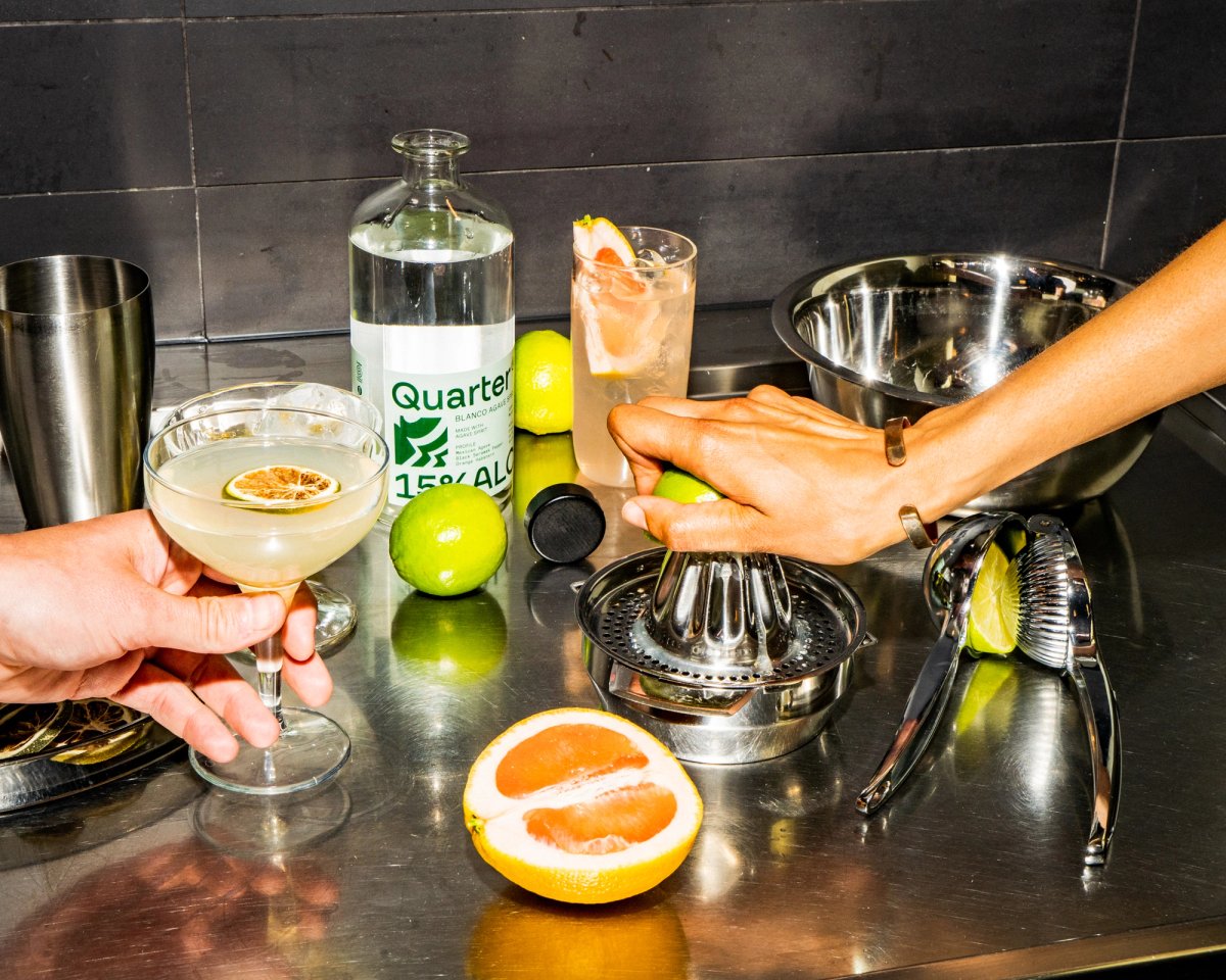 A hand squeezes a citrus wedge into a glass cocktail, garnished with a slice of orange. A bottle of Quarter 15% Alc stands nearby, along with additional citrus fruits and cocktail-making tools.