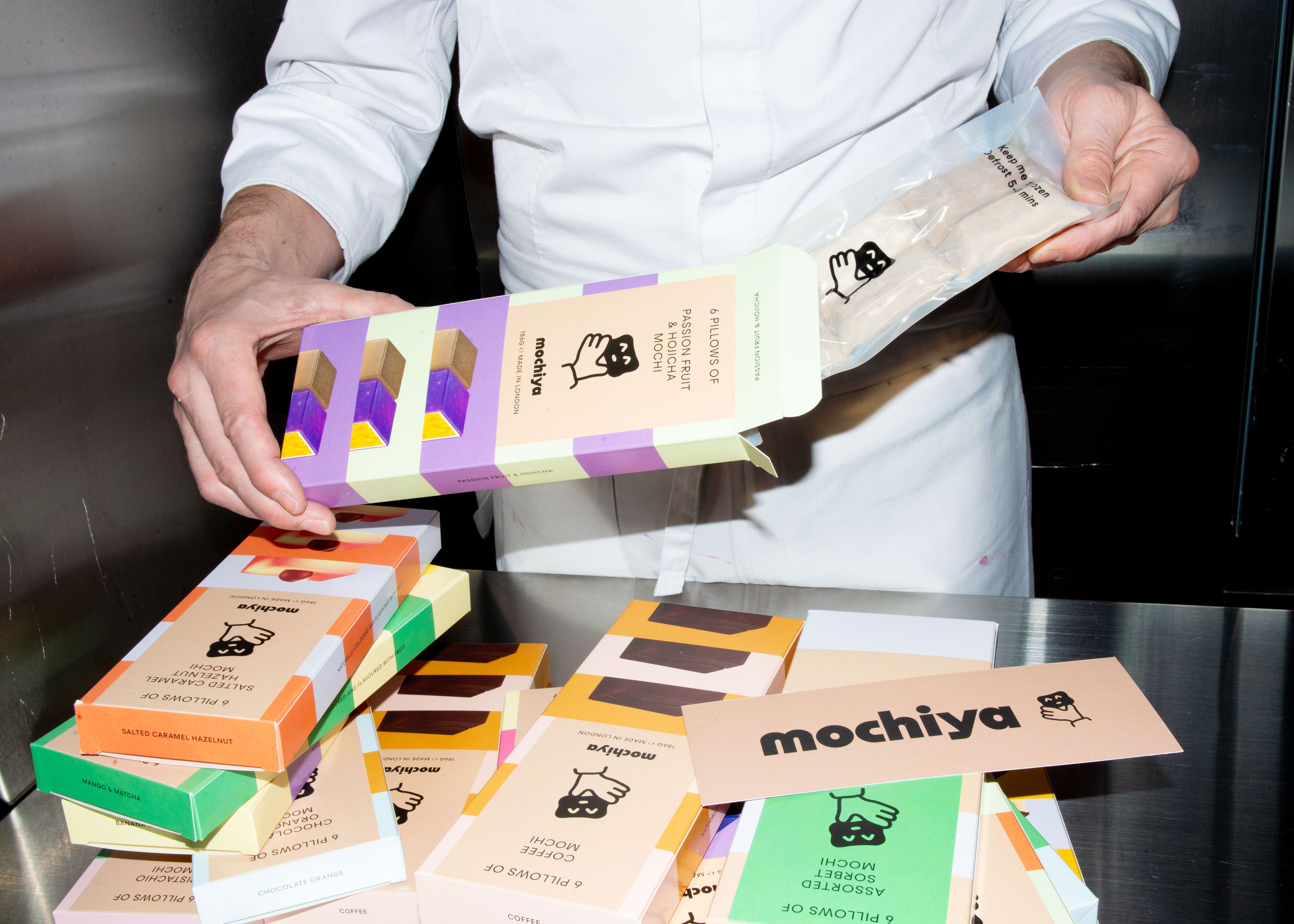 A person holds a box of Mochiya Pills of Fruit and Passion Fruit, a salted caramel hazelnut flavor, alongside several boxes of Mochiya Mochi Sorbet.