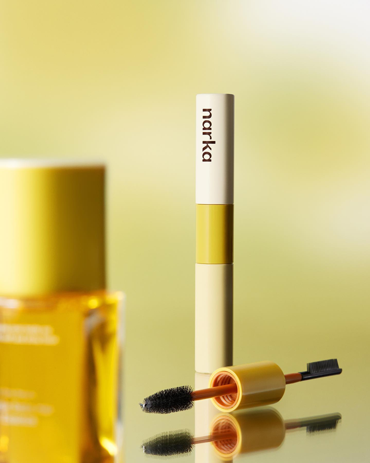 A Marka mascara tube and a mascara brush with black bristles are displayed against a yellow-green gradient background.