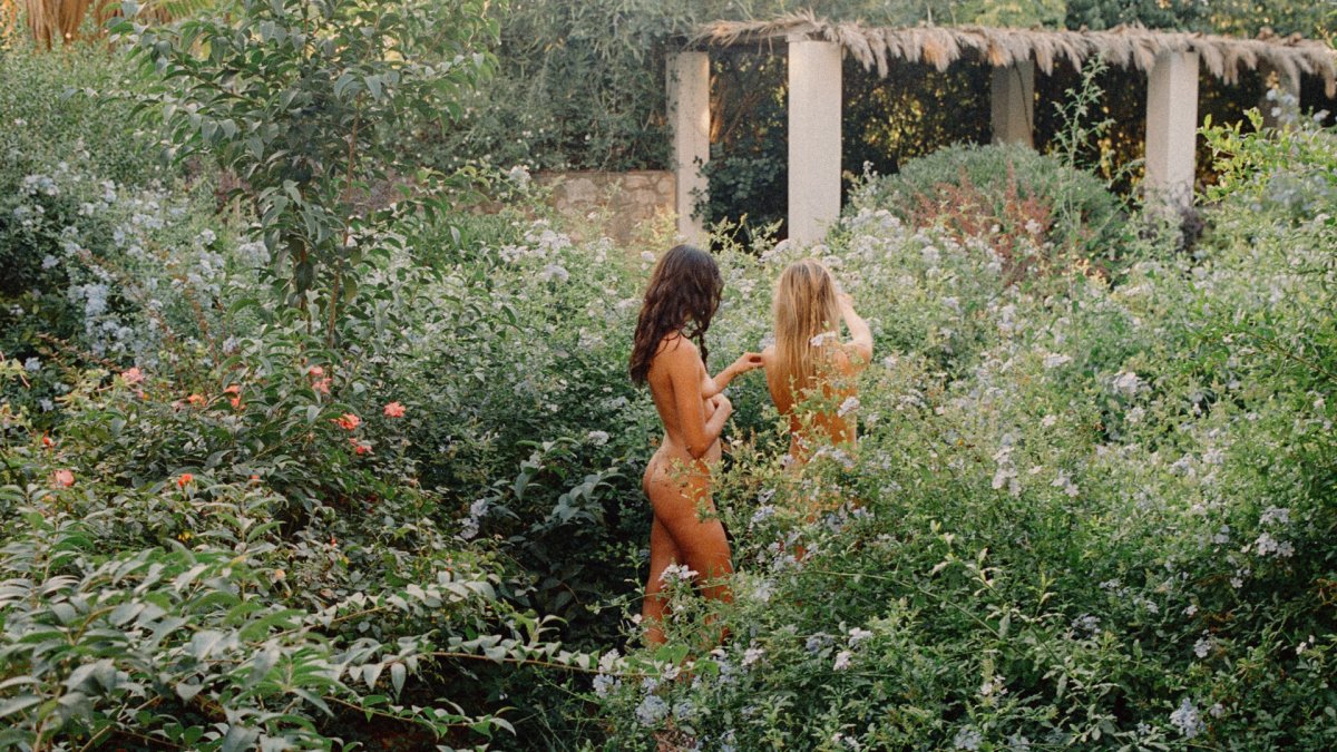 Two individuals in light-colored clothing stand amidst dense vegetation in a garden setting.  A thatched structure is partially visible in the background.