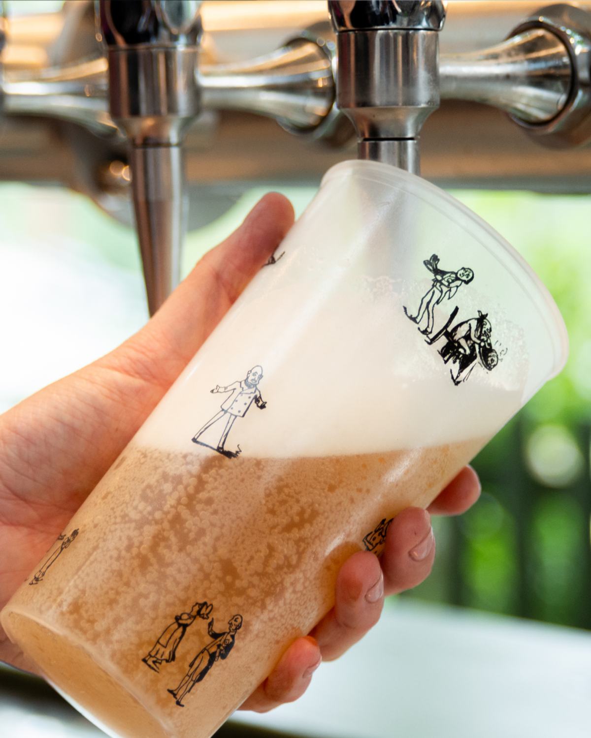 A hand holds a plastic cup filled with a light-colored beverage, pouring from a tap. The cup features black drawings of people.