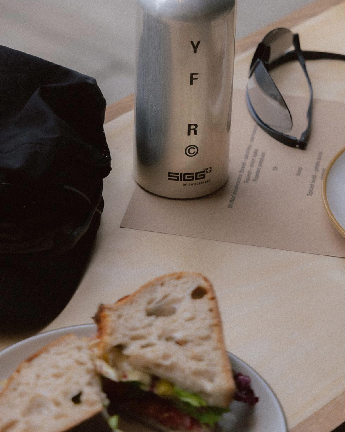 A silver SIGG+ water bottle, a black cap, a pair of eyeglasses, and a partially eaten sandwich are arranged on a light wooden surface. A piece of paper with text is also visible.