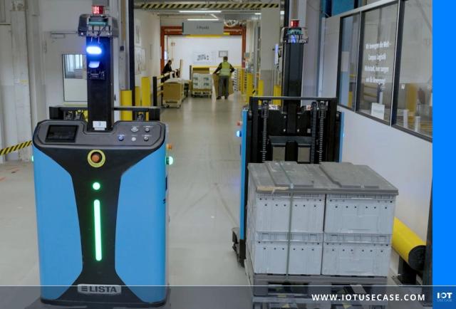 Zwei blaue fahrerlose Transportfahrzeuge in einer Werkshalle von Schaeffler: links ein AGV mit grünem Leuchtstreifen, rechts ein Gabel-AGV, das gestapelte graue Behälter auf einer Palette transportiert; im Hintergrund ein Mitarbeiter in Warnweste.