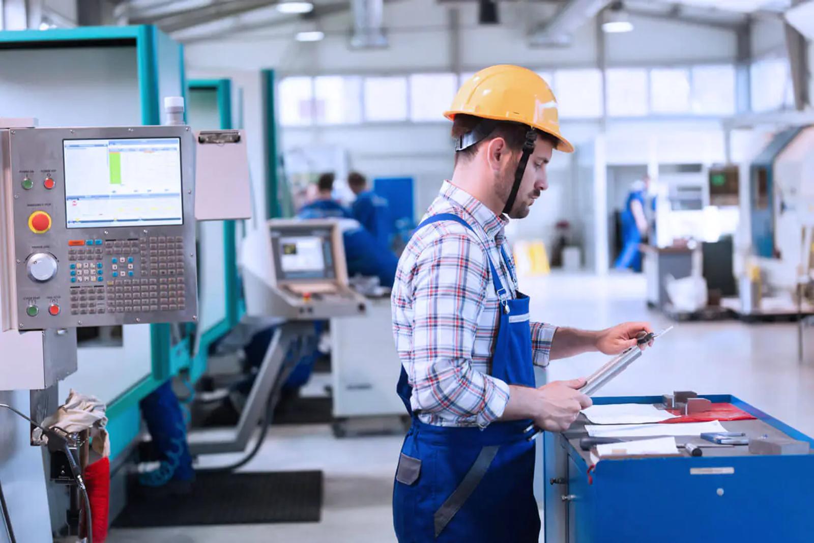Produktionsarbeiter mit gelbem Helm prüft Unterlagen neben CNC-Maschine