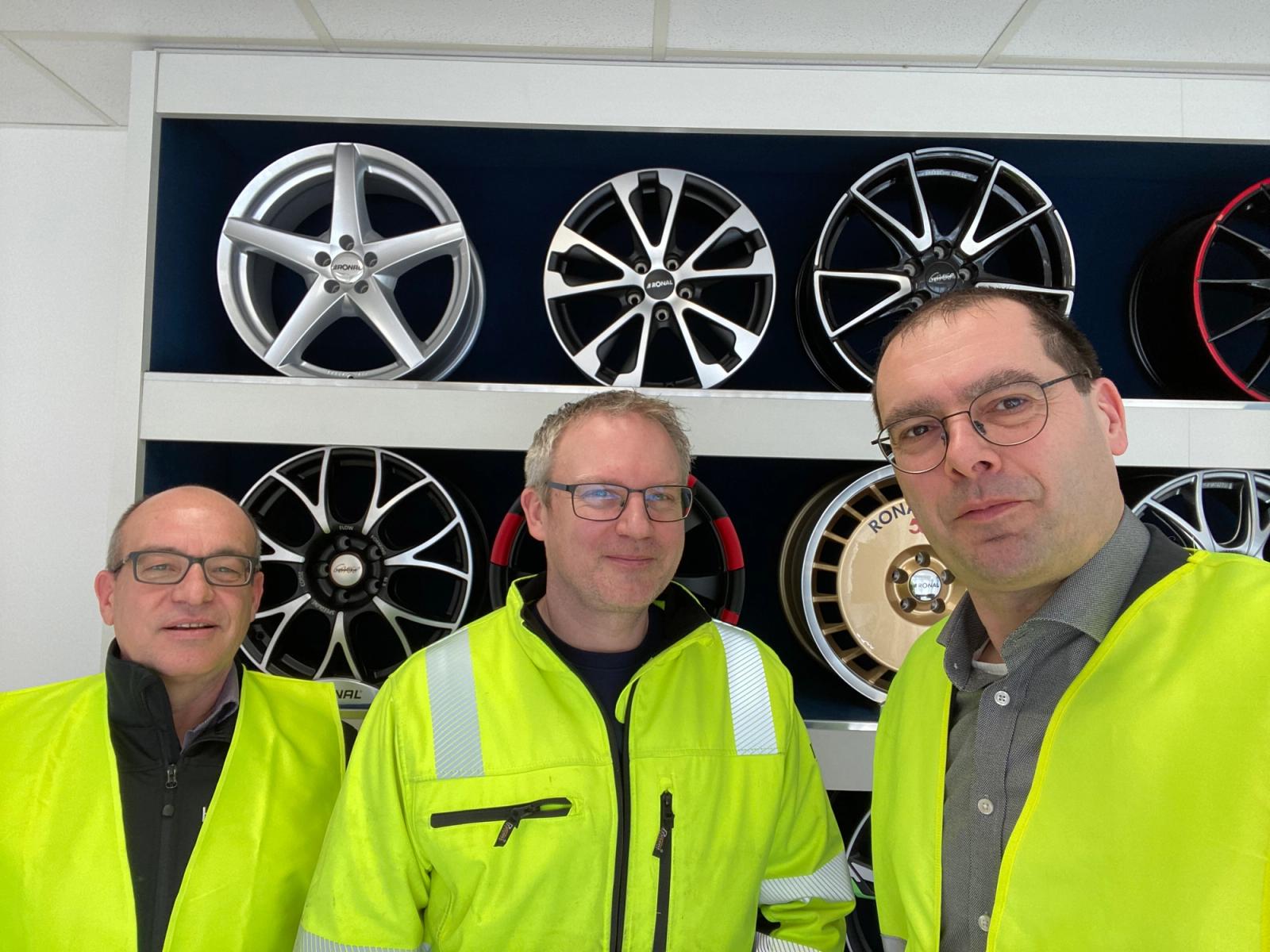 Three employees in yellow safety vests in front of wheel rim display wall at compressed air leak project