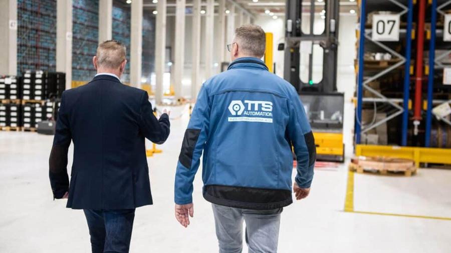 Zwei Männer, einer in TTS-Jacke, gehen durch eine Logistikhalle