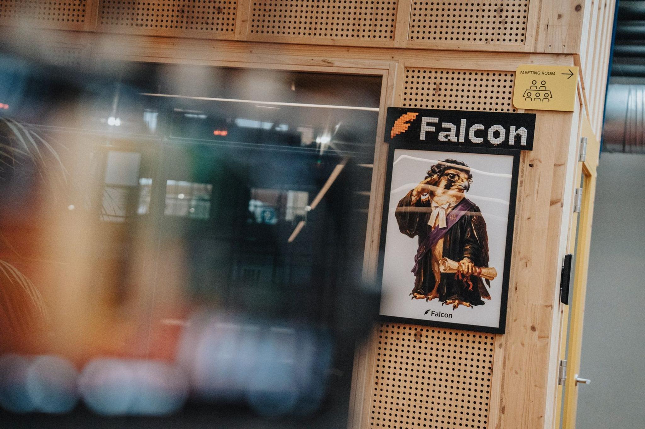 Achter de schermen: Falcon-logo en mascotteposter aan een vergaderruimte, met de camera wazig op de voorgrond