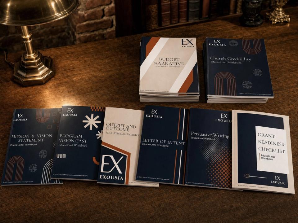 The Exousia educational workbooks laid out on an oak library table, several fanned in an arc and the rest in short stacks, under the warm light of a brass lamp.