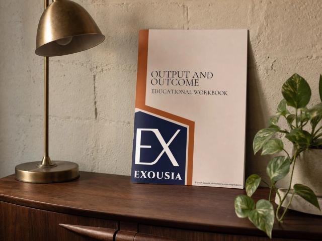 The Output and Outcome workbook leaning against a plaster wall on a walnut credenza, beside a brass desk lamp and a trailing pothos.