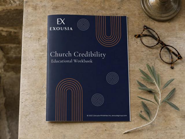 The Church Credibility workbook seen from above on a worn stone-coloured table, with tortoiseshell reading glasses and a sprig of olive.