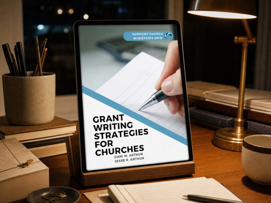 The cover of Grant Writing Strategies for Churches displayed on a tablet propped in a wooden stand on a desk at night, lit by a brass lamp.